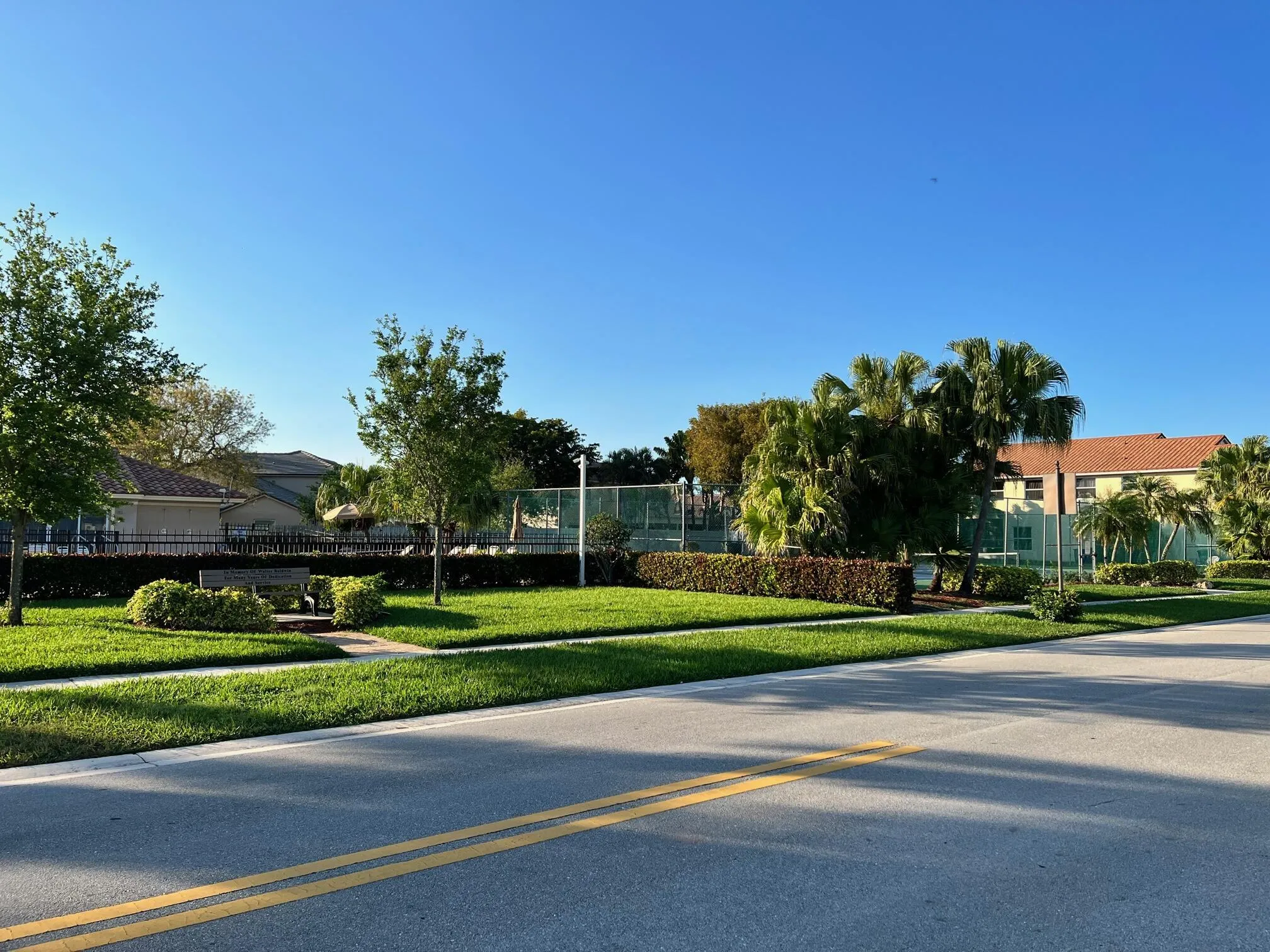 Community green space featuring a manicured lawn, mature trees, and a paved pathway leading to a bench