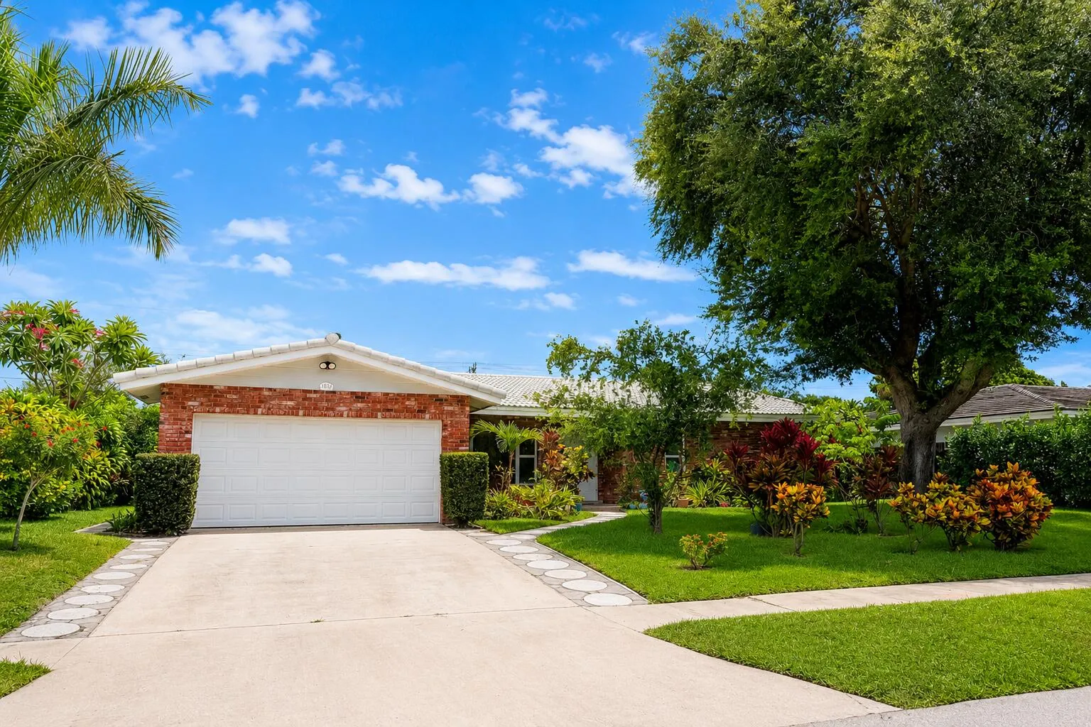 Red brick facade home featuring a white garage door, concrete driveway with decorative stepping stones, a lush green lawn, and mature trees and shrubs