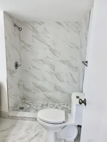 Bathroom featuring a walk-in shower with large format marble-look tile walls, a hexagonal mosaic tile shower pan, and a toilet