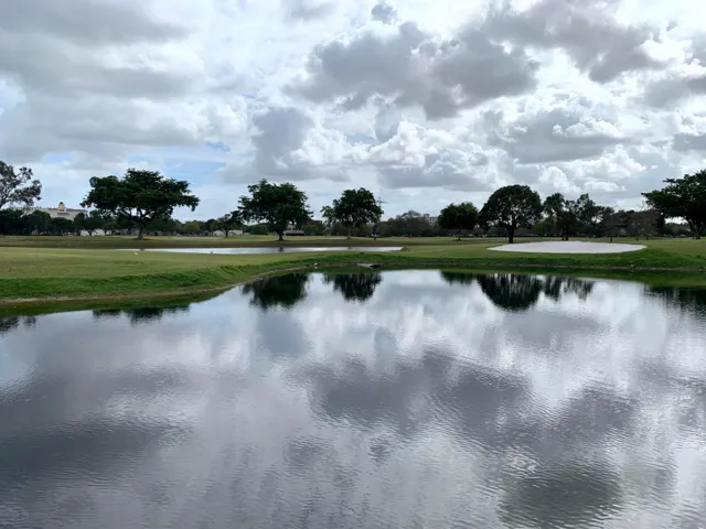 Expansive golf course frontage featuring a water hazard, manicured green space, mature trees, and a distant sand trap