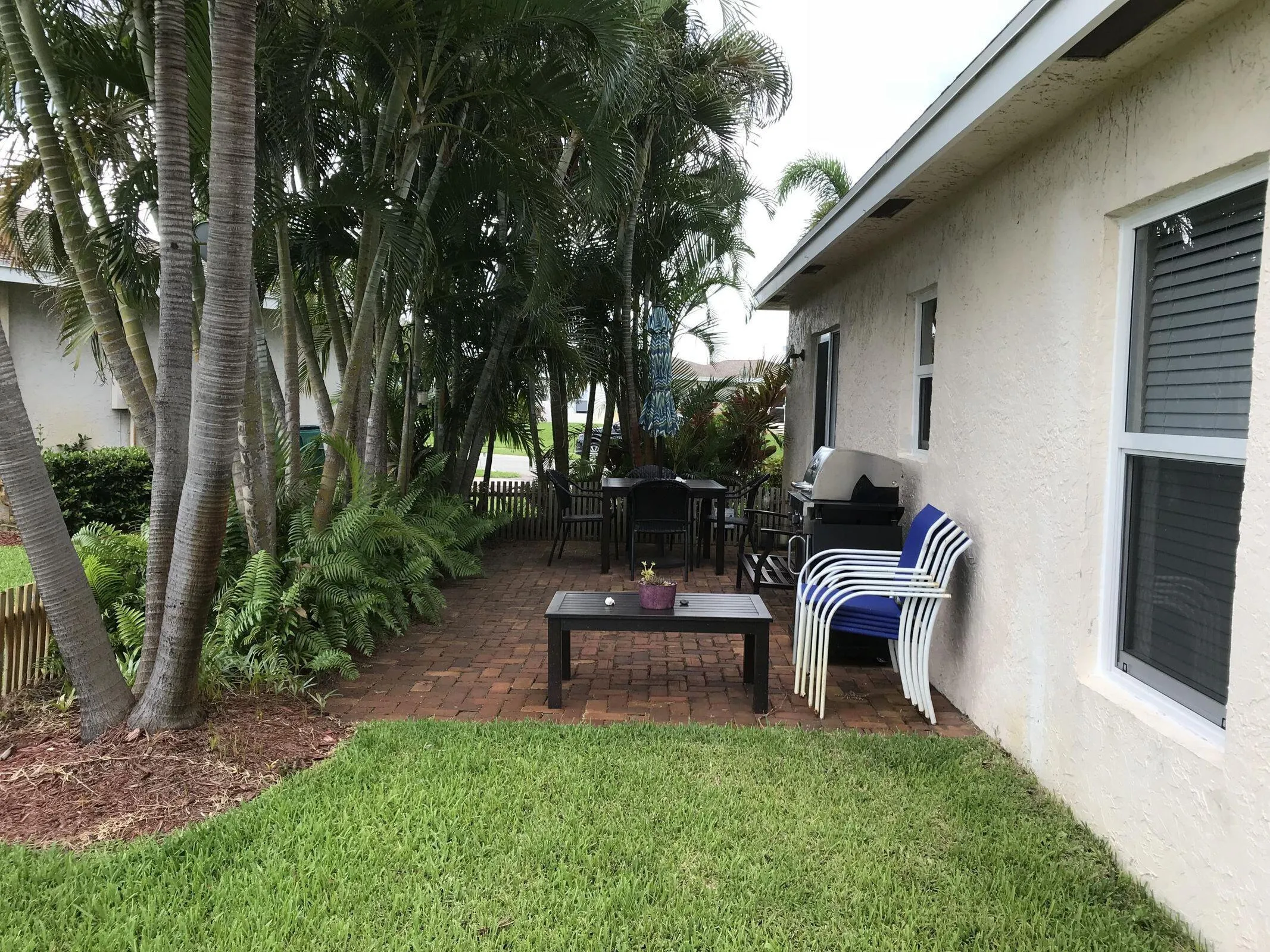 Expansive brick paver patio featuring a partial privacy fence, lush mature palm trees, tropical plantings, and a manicured lawn