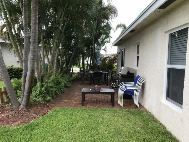 Expansive brick paver patio featuring a partial privacy fence, lush mature palm trees, tropical plantings, and a manicured lawn