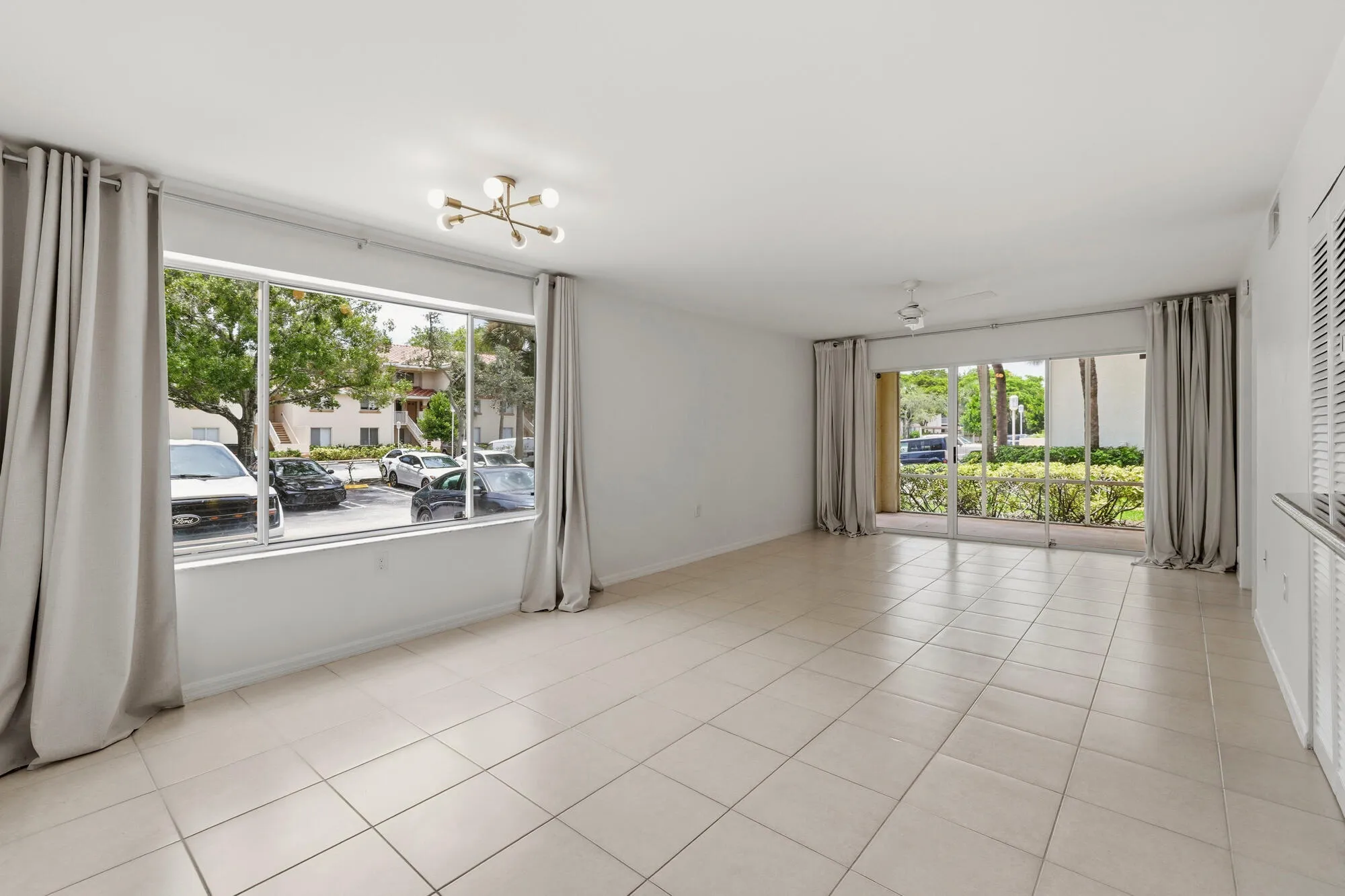 Spacious living area featuring neutral tile flooring, a wide window with light-filtering drapes, and a contemporary ceiling fixture