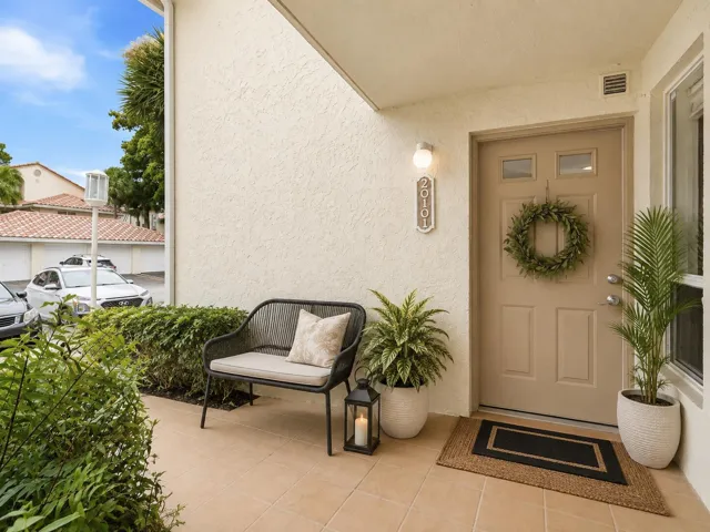 Welcoming South Florida condo entrance with a front door, outdoor bench, potted tropical plants, lantern, wreath, and layered welcome mat.