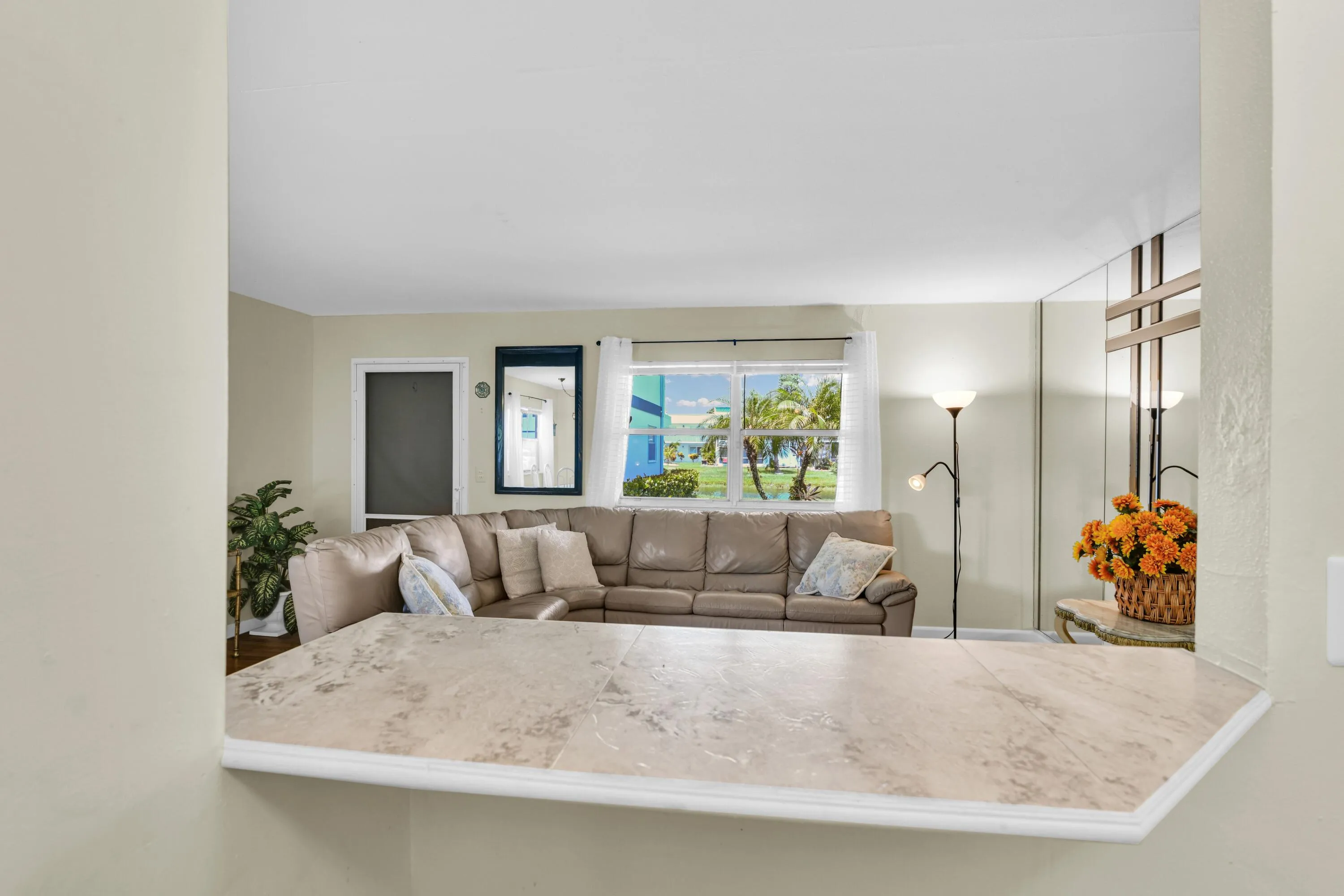 Kitchen pass-through featuring a light-toned countertop, opening to a living area with large windows, wall-mounted mirror, and decorative wall paneling