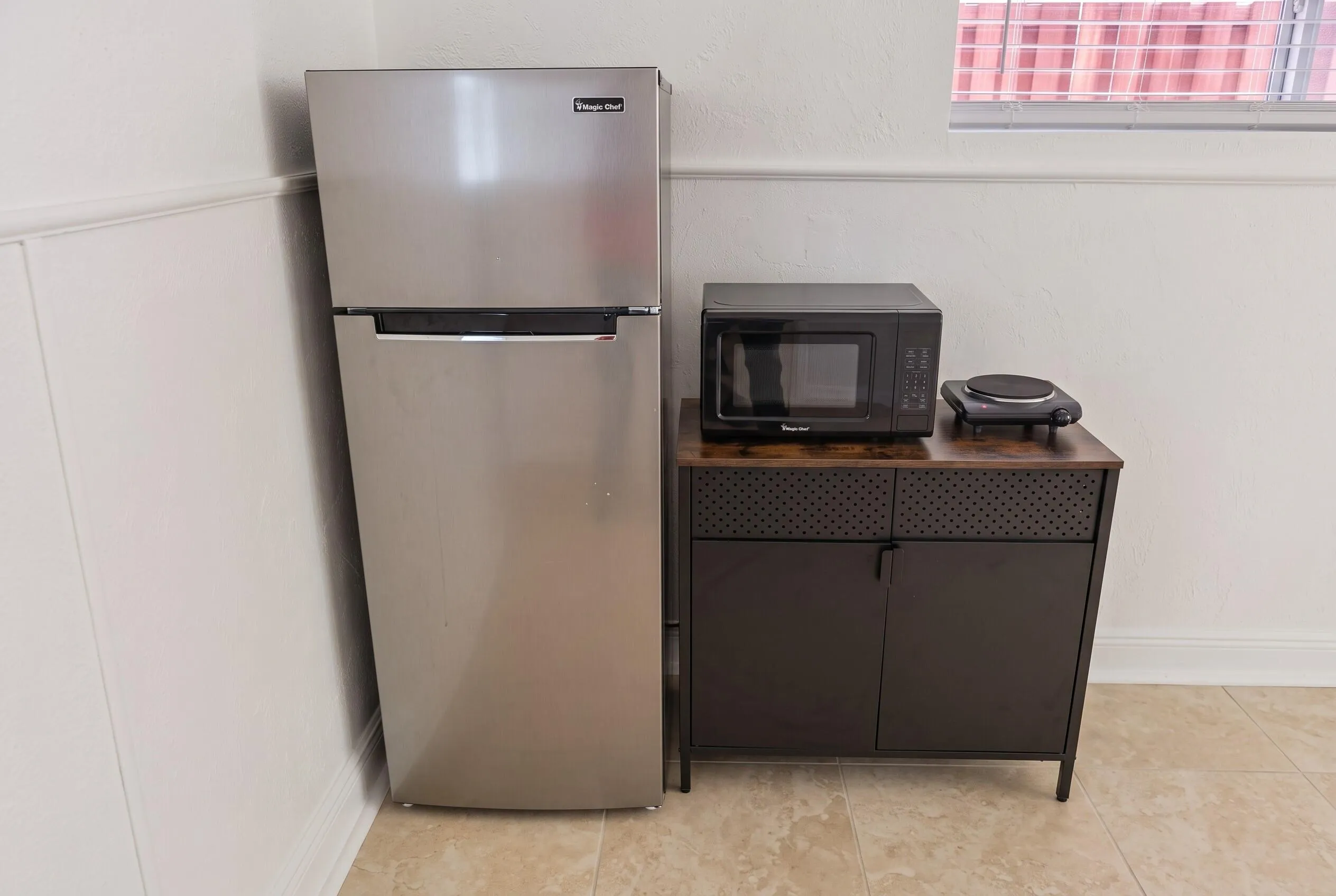 Compact kitchen area featuring a stainless steel refrigerator, microwave, and single burner hot plate, accompanied by a two-door storage cabinet with textured drawer fronts and a wood-finish top