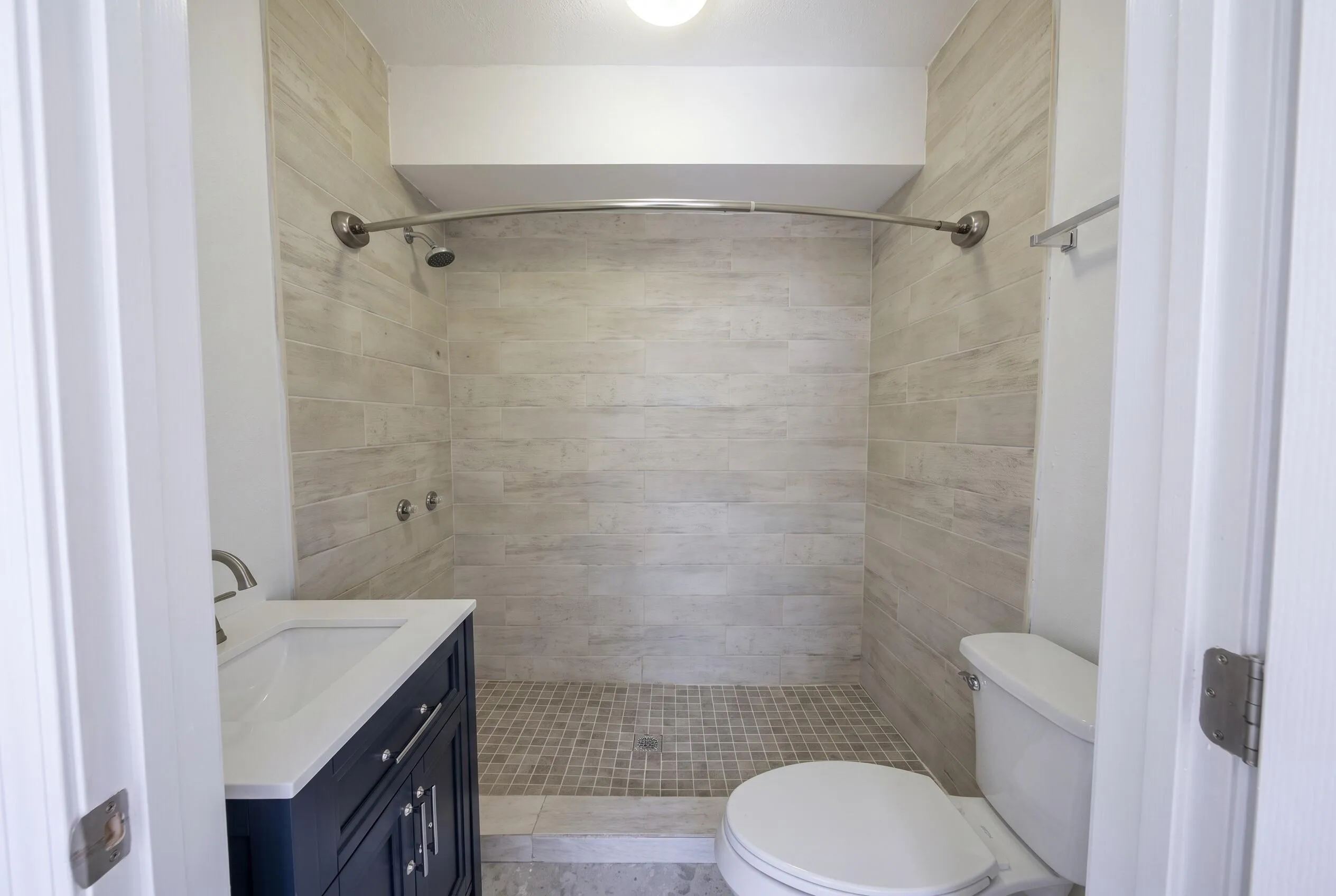 Bathroom featuring a tiled shower stall with a curved shower rod, a dark blue vanity with a white countertop and integrated sink, a toilet, and light-toned wall and floor tiling
