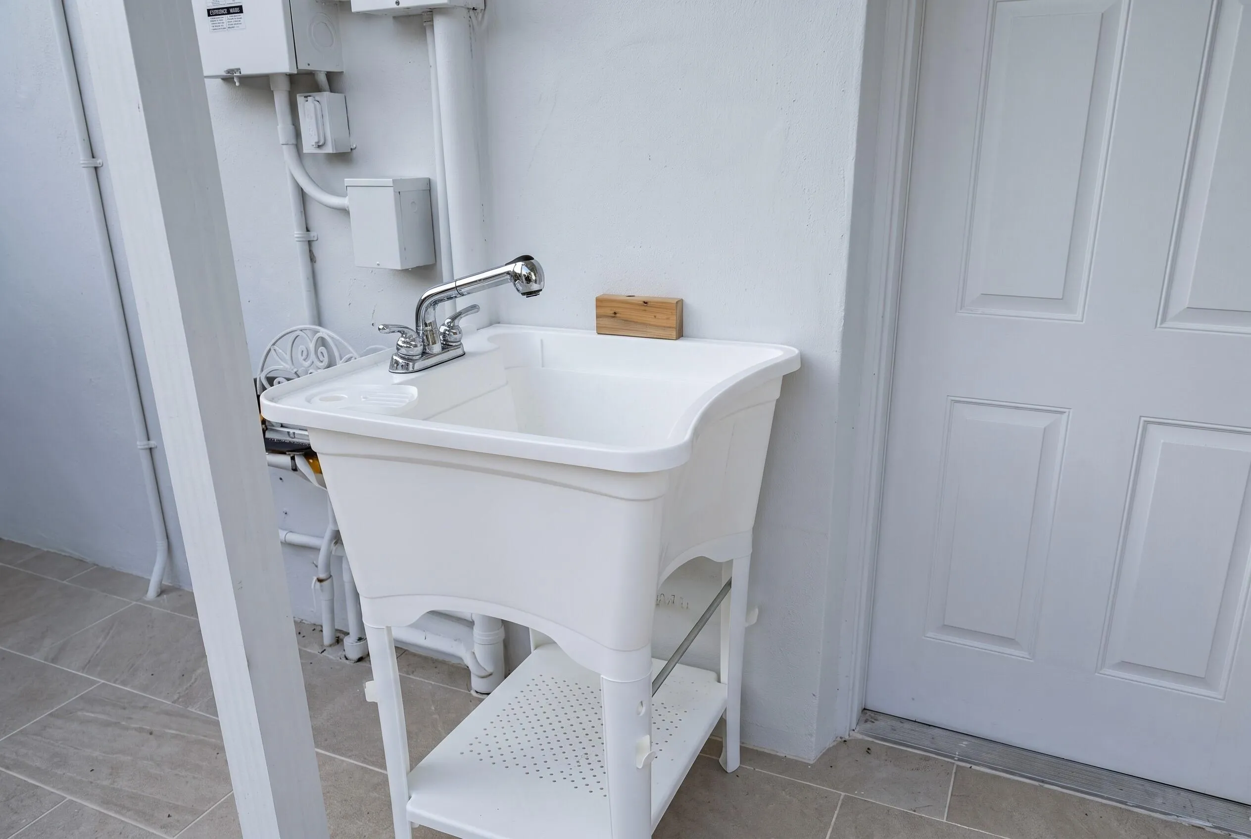 Utility sink with chrome faucet, featuring a lower storage shelf, set against a white wall with visible utility boxes and conduit