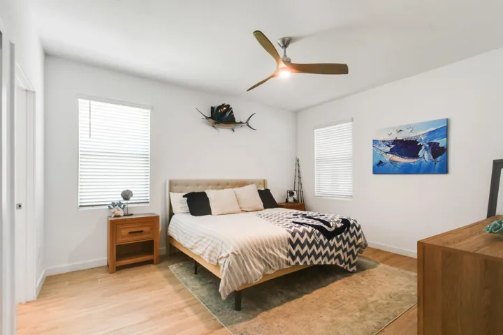 Spacious bedroom featuring light wood-plank tile flooring, ample natural light from two windows with blinds, a ceiling fan with integrated lighting, and white walls