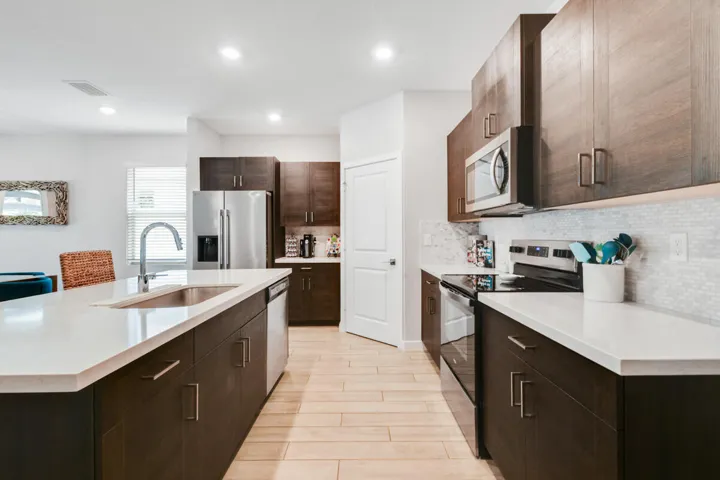 Modern kitchen featuring a large island with an undermount sink, stainless steel appliances, dark wood-finish cabinetry, light quartz countertops, and a soft mosaic tile backsplash