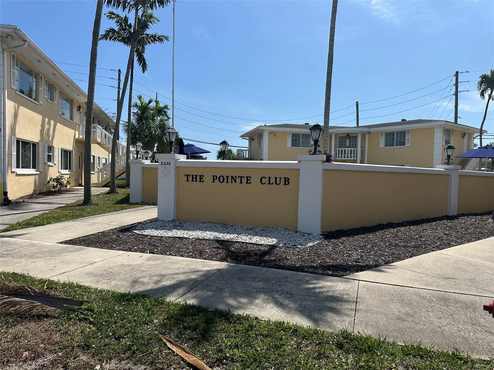 "The Pointe Club" entrance monument featuring exterior lighting, manicured landscaping with dark and light rock accents, and a paved sidewalk