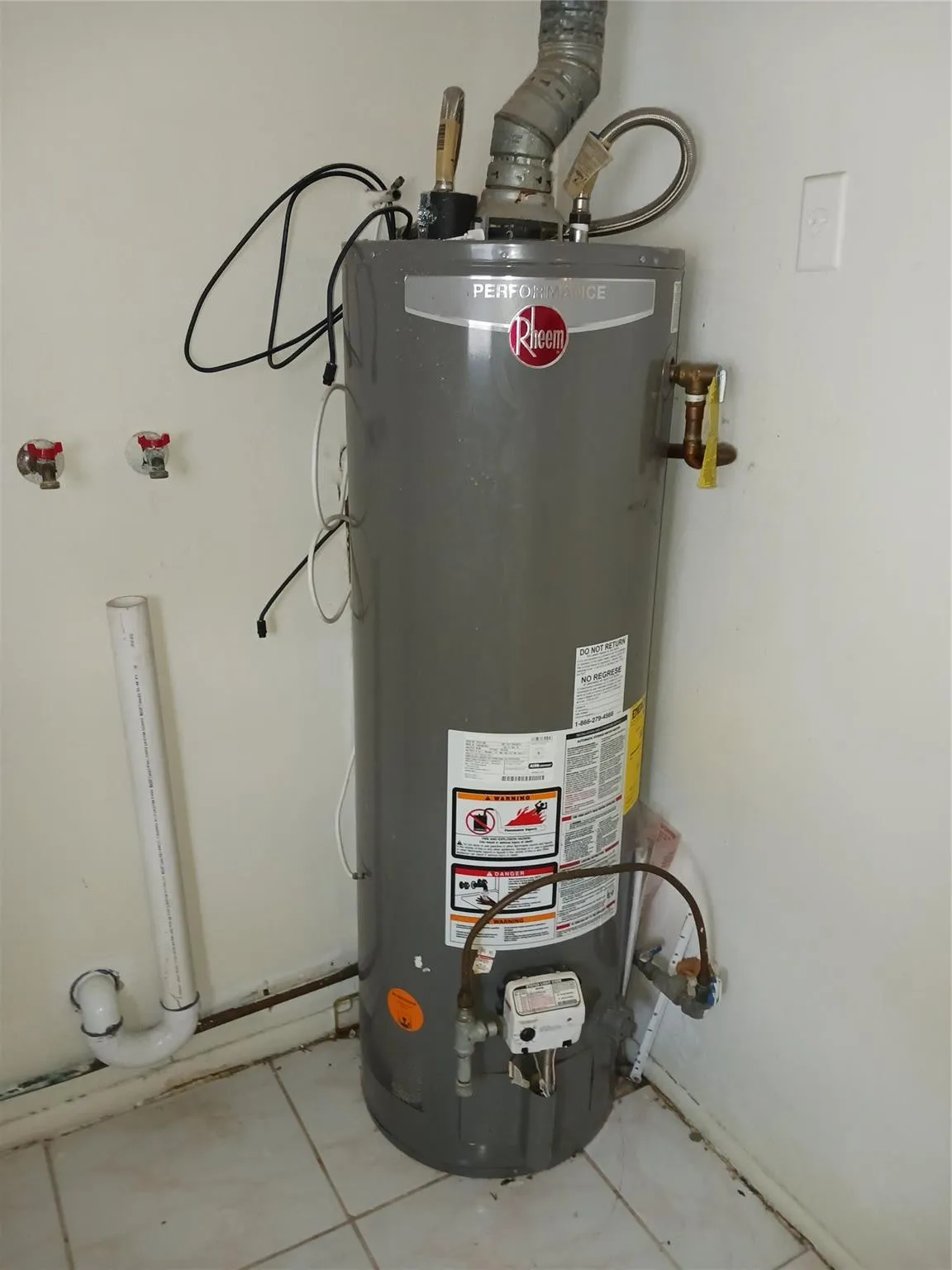 Water heater installation with visible utility connections and tiled flooring