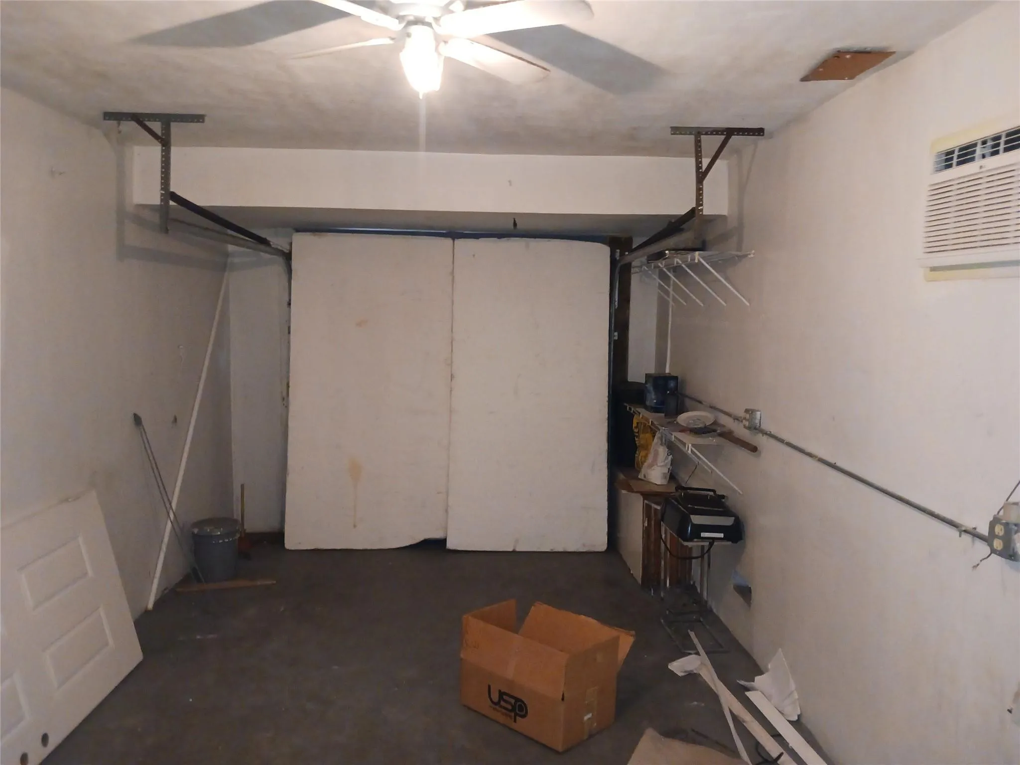 Garage interior featuring a ceiling fan with integrated lighting, a wall-mounted air conditioning unit, and exposed ceiling joists