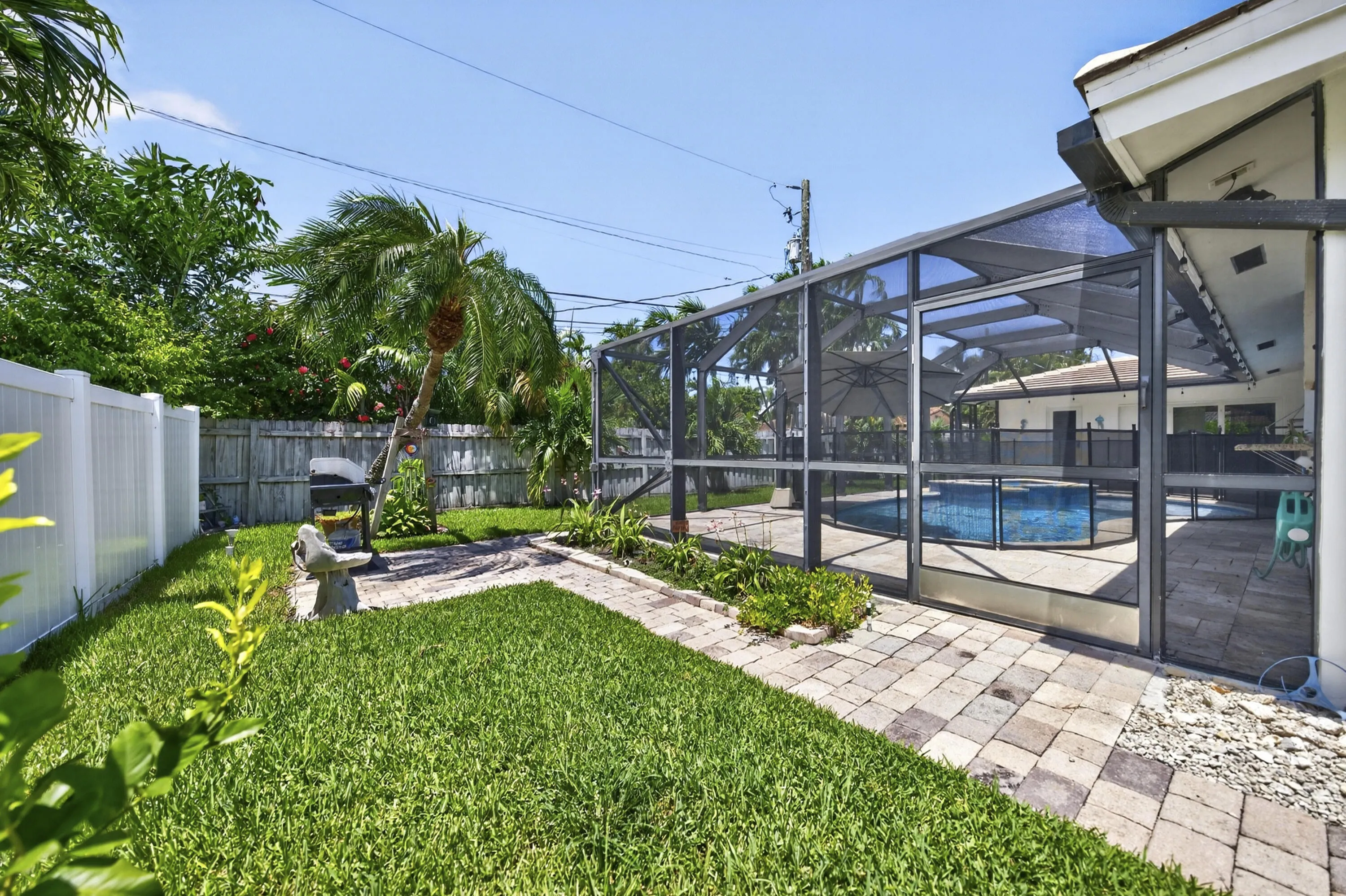 Backyard featuring a screened-in pool enclosure, paver walkway, mature palm trees, and a white privacy fence