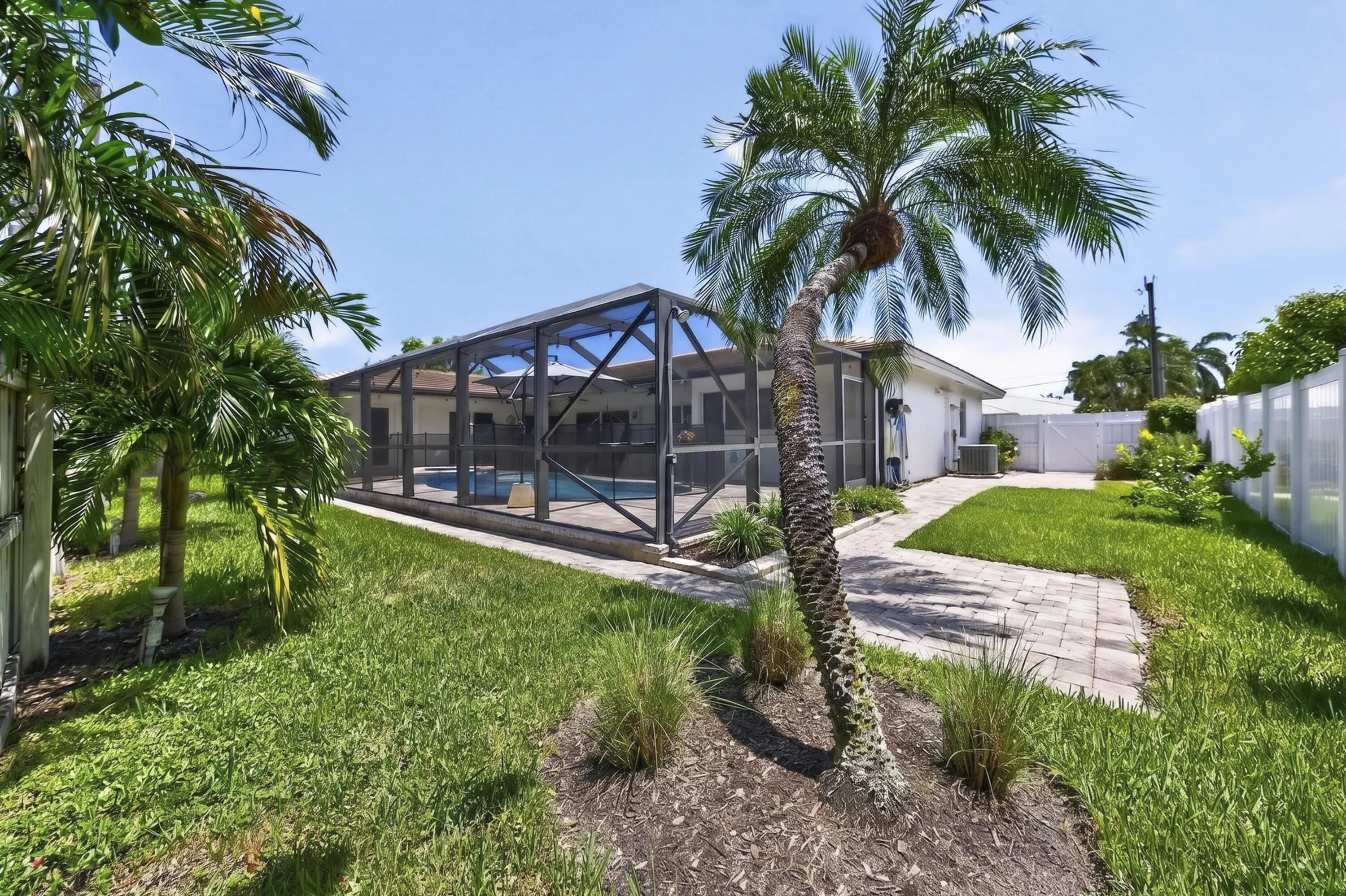 Backyard featuring a screened-in pool enclosure, paver walkway, mature palm trees, and a white privacy fence