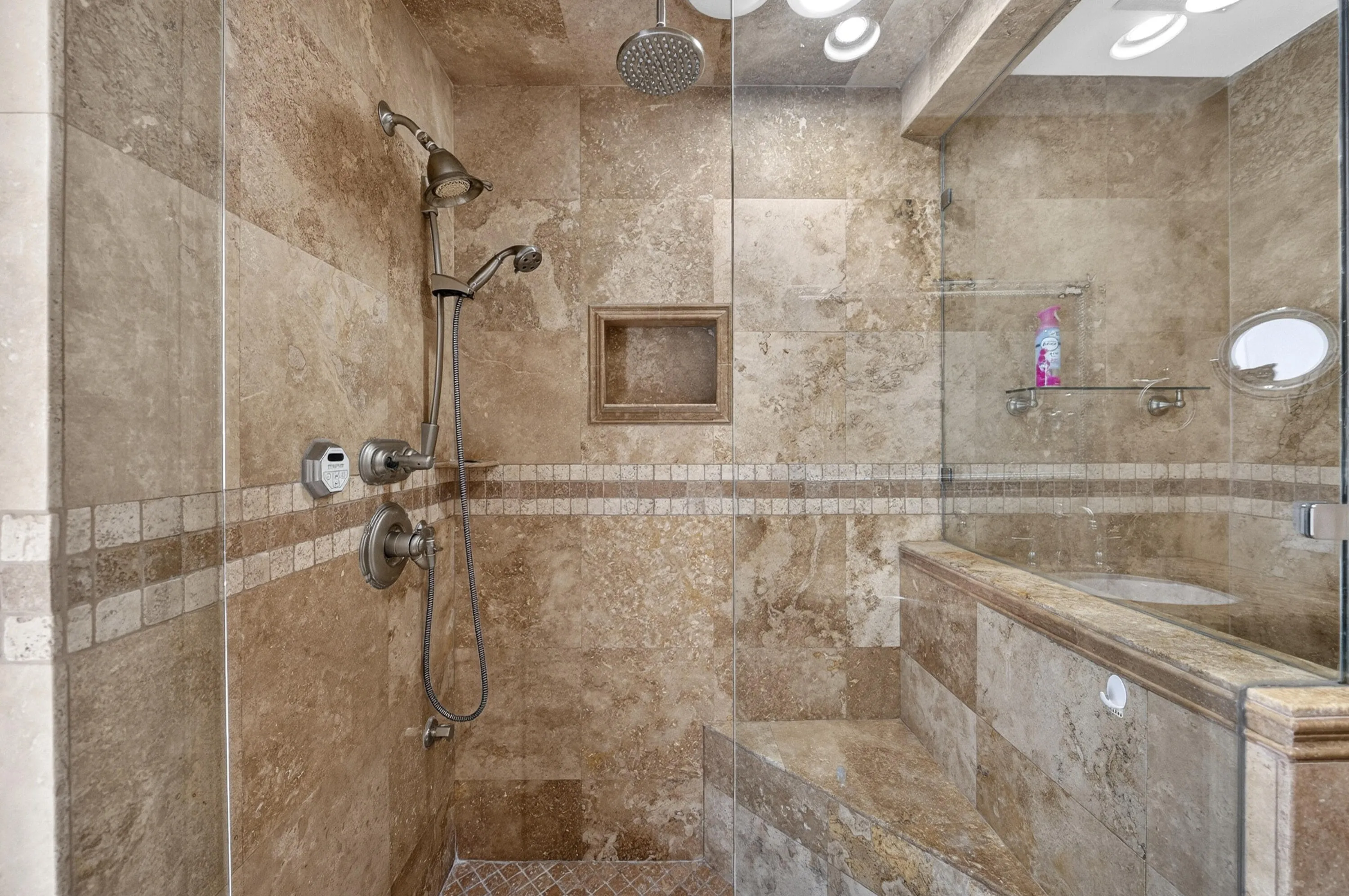 Expansive shower featuring stone-look tile, a built-in bench, glass enclosure, recessed wall niche, and multiple shower heads