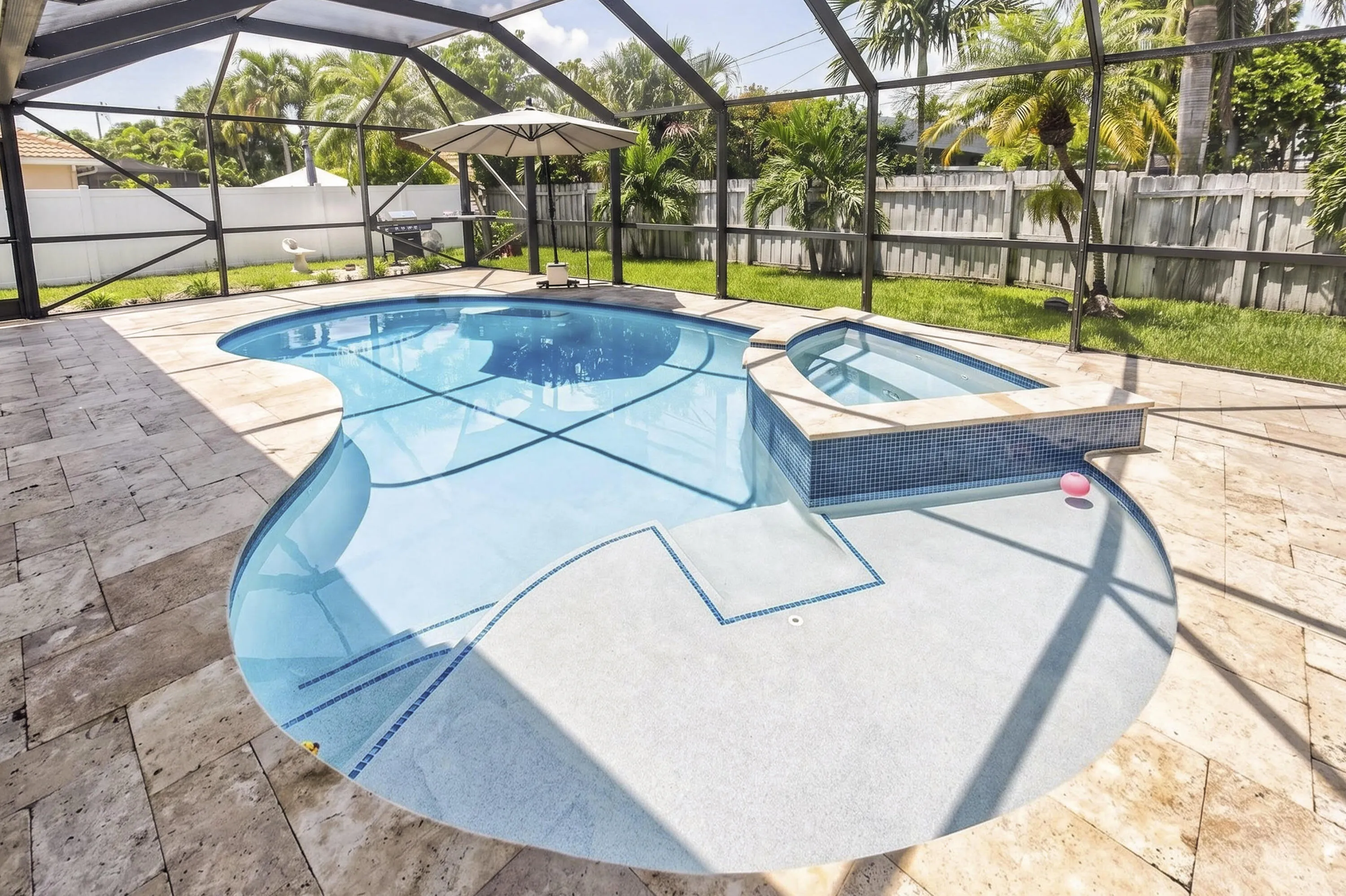 Screened outdoor area featuring a freeform swimming pool with integrated spa, light-colored travertine tile decking, and a fenced perimeter