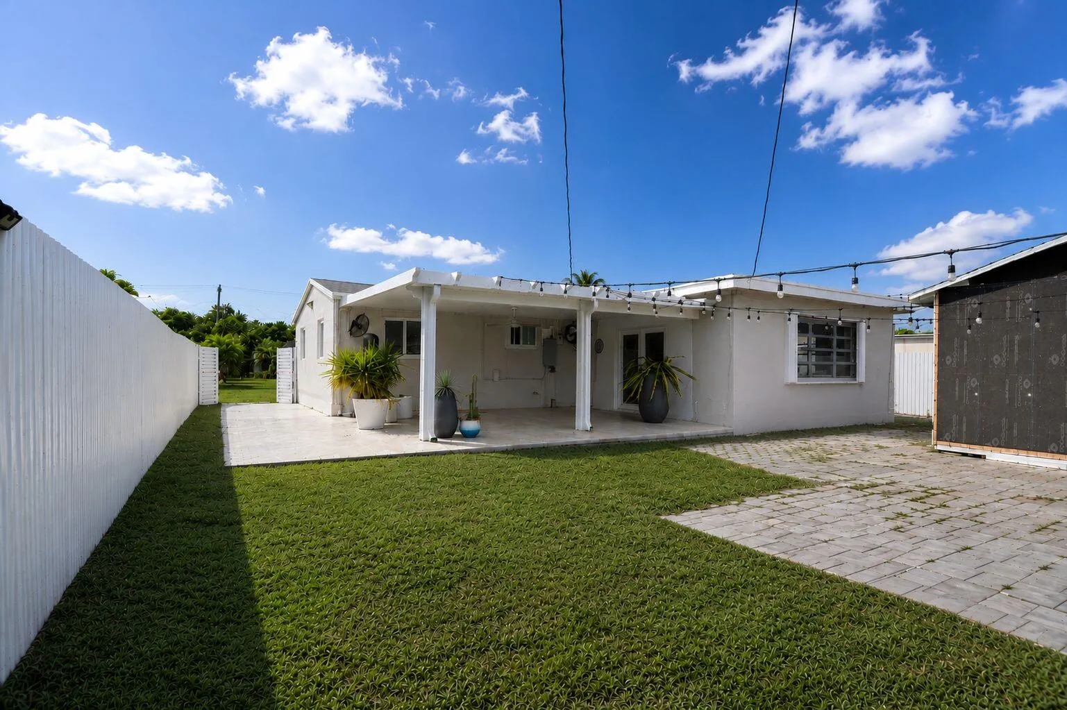 Expansive backyard featuring a covered patio, a manicured lawn, a white privacy fence, and a paver stone patio area