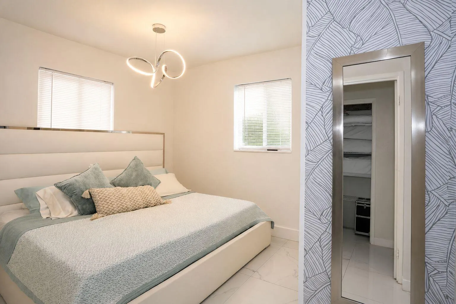Bright bedroom featuring two windows with blinds, a contemporary chandelier, white walls, and a patterned accent wall