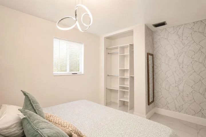Modern overhead light fixture, window with blinds, open closet system with shelving and hanging rods, patterned accent wall, and tiled flooring