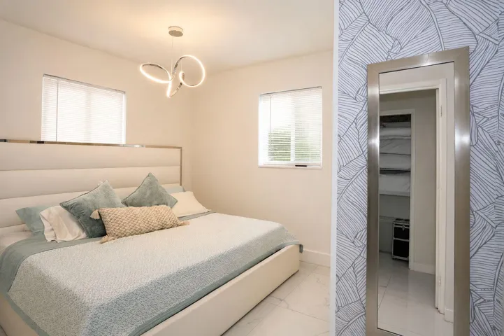 Bright bedroom featuring two windows with blinds, a contemporary chandelier, white walls, and a patterned accent wall