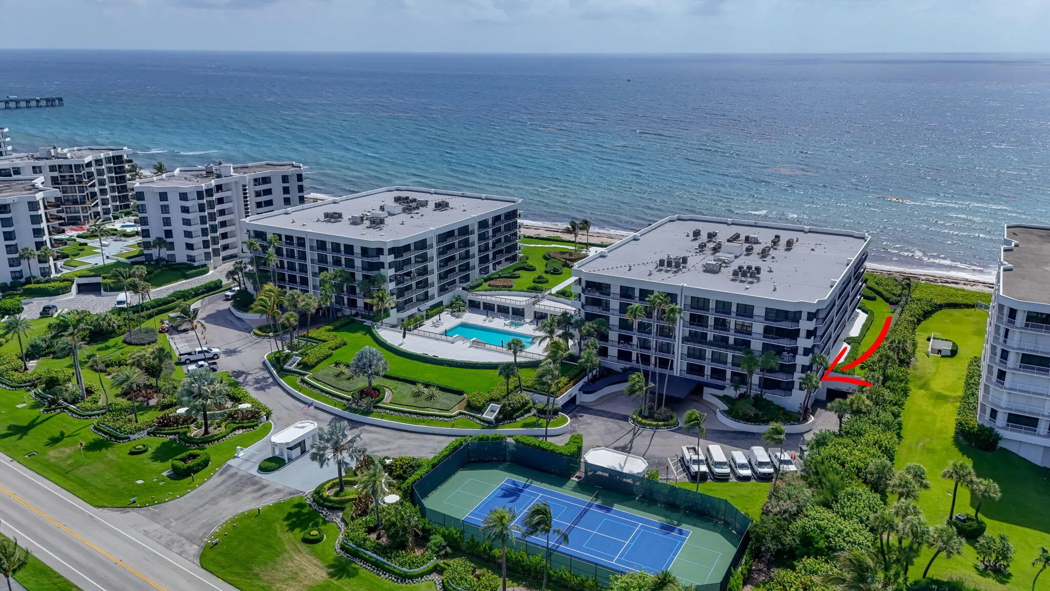 Oceanfront condominium building featuring multiple balconies, a ground-level swimming pool with a paved deck, and an adjacent tennis court