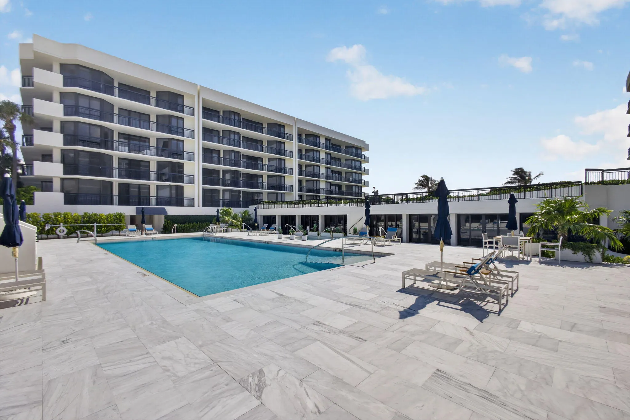 Expansive pool deck featuring a rectangular swimming pool, light-toned stone tile flooring, and a multi-story building with private balconies