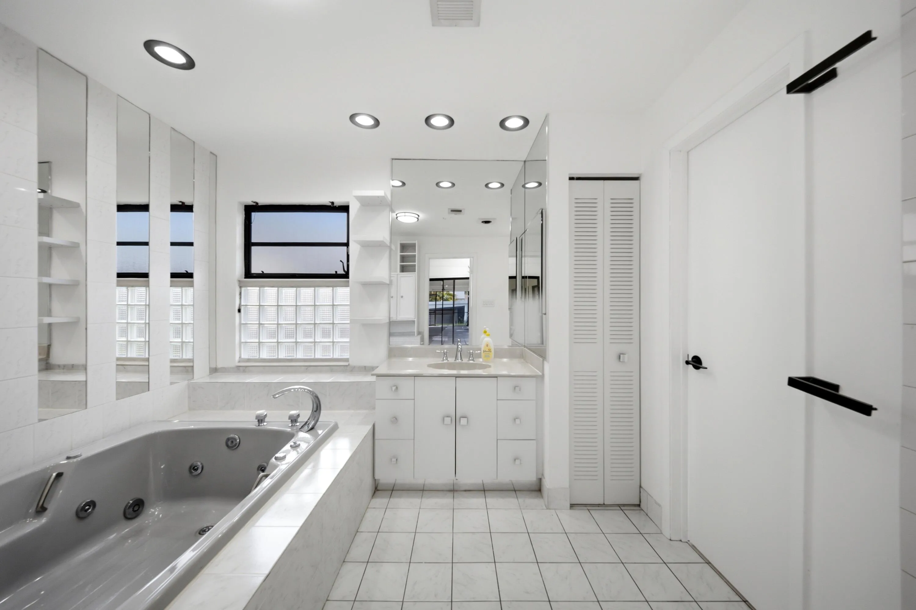 Bathroom featuring a jetted tub, wall-mounted mirrored vanity, built-in shelving, glass block window, and recessed lighting