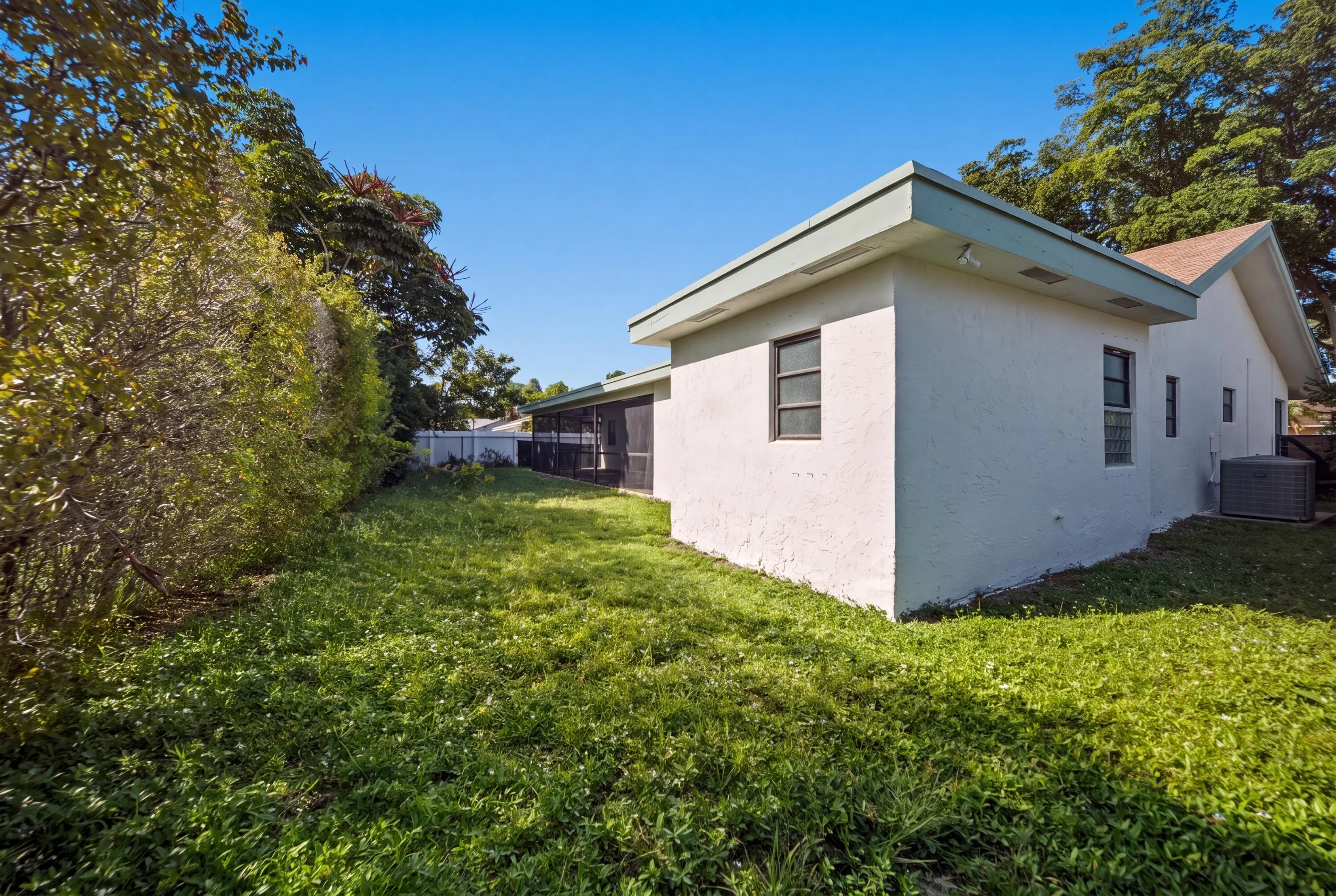 Expansive grassy yard with mature perimeter foliage, a stucco exterior, multiple window openings, and a screened enclosure