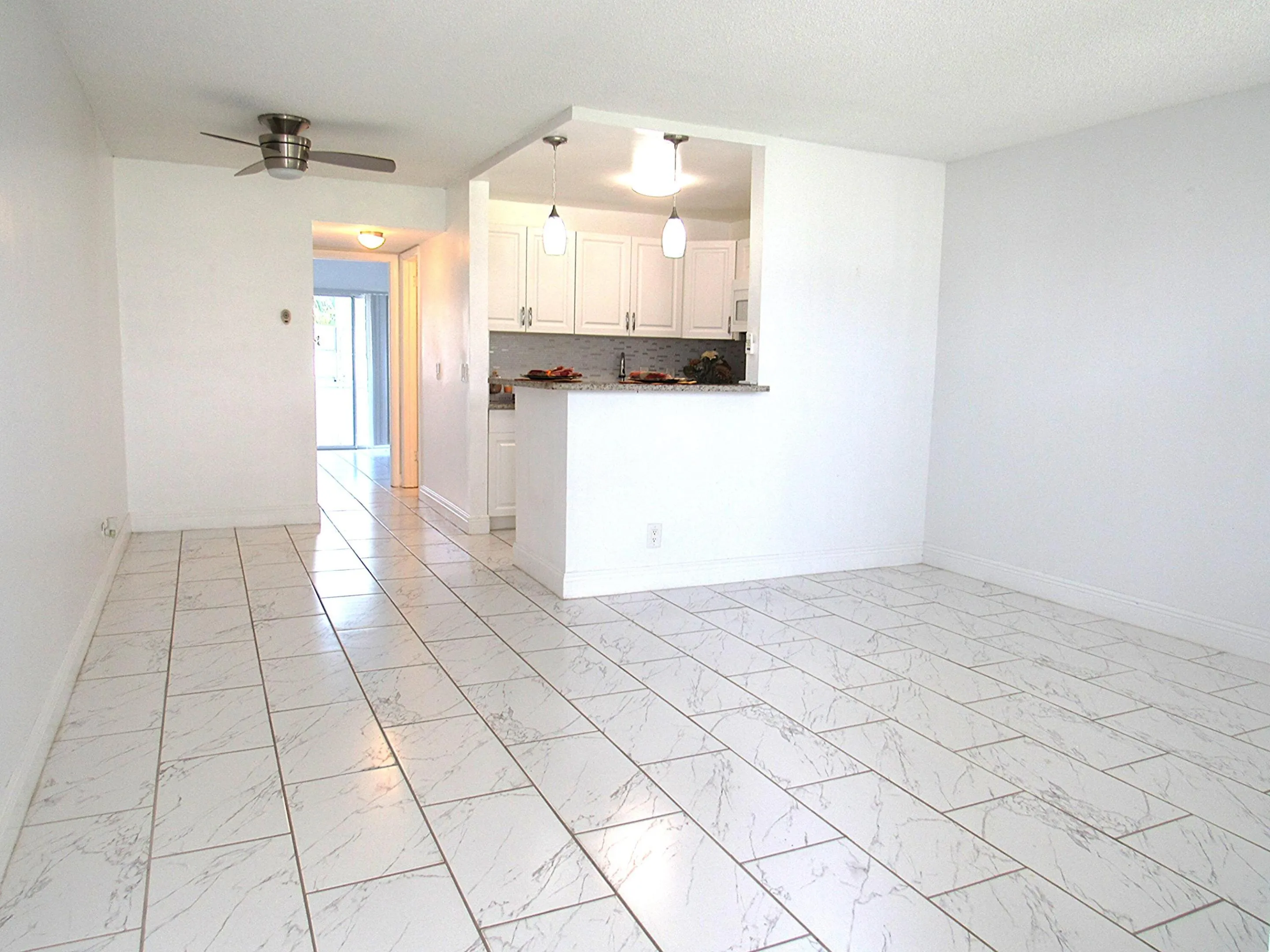 Spacious interior featuring white tile flooring, crisp white walls, a pass-through kitchen opening, a contemporary ceiling fan.