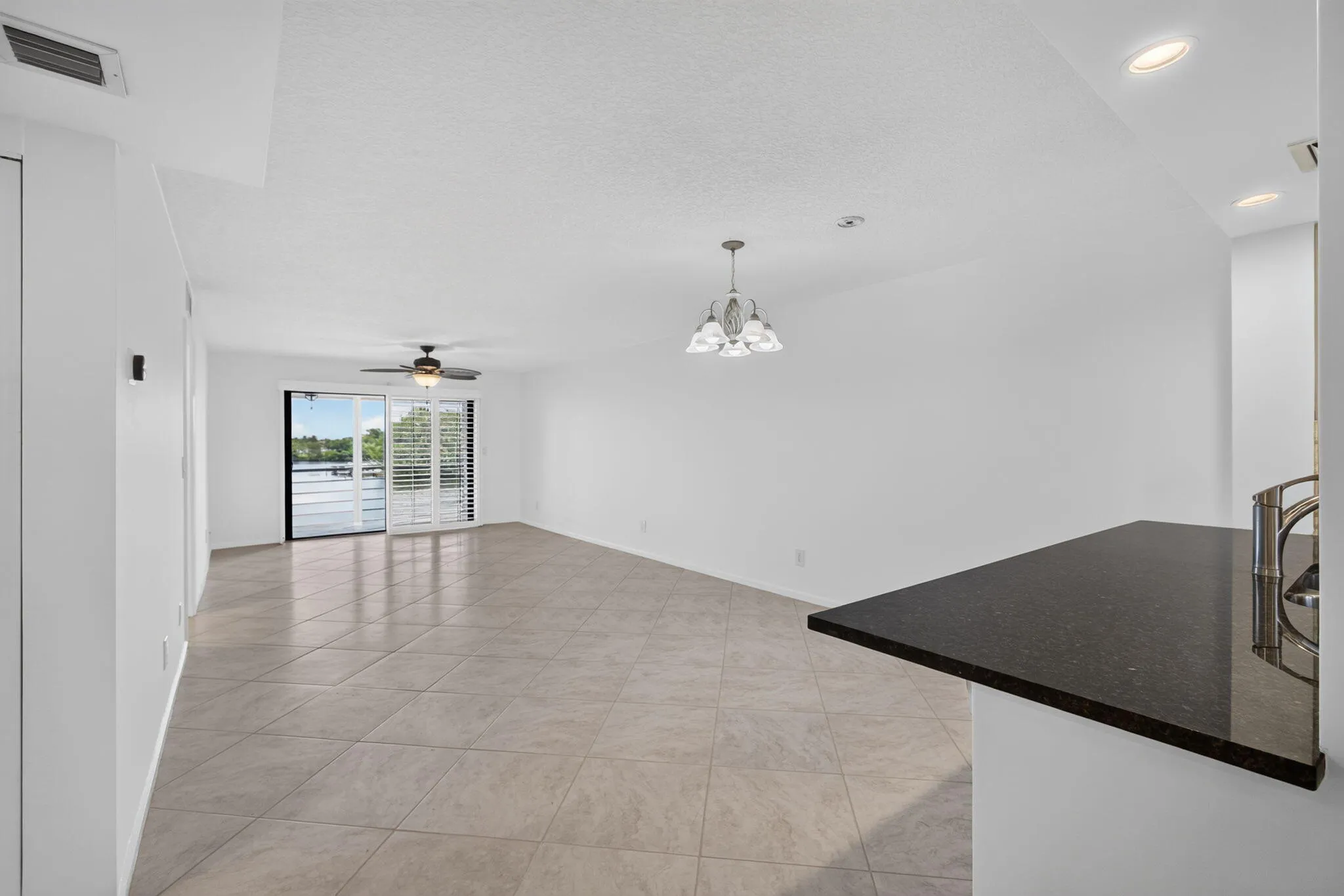 Spacious room featuring tile flooring, sliding glass doors with water scenery, plantation shutters, a ceiling fan, and a four-light chandelier