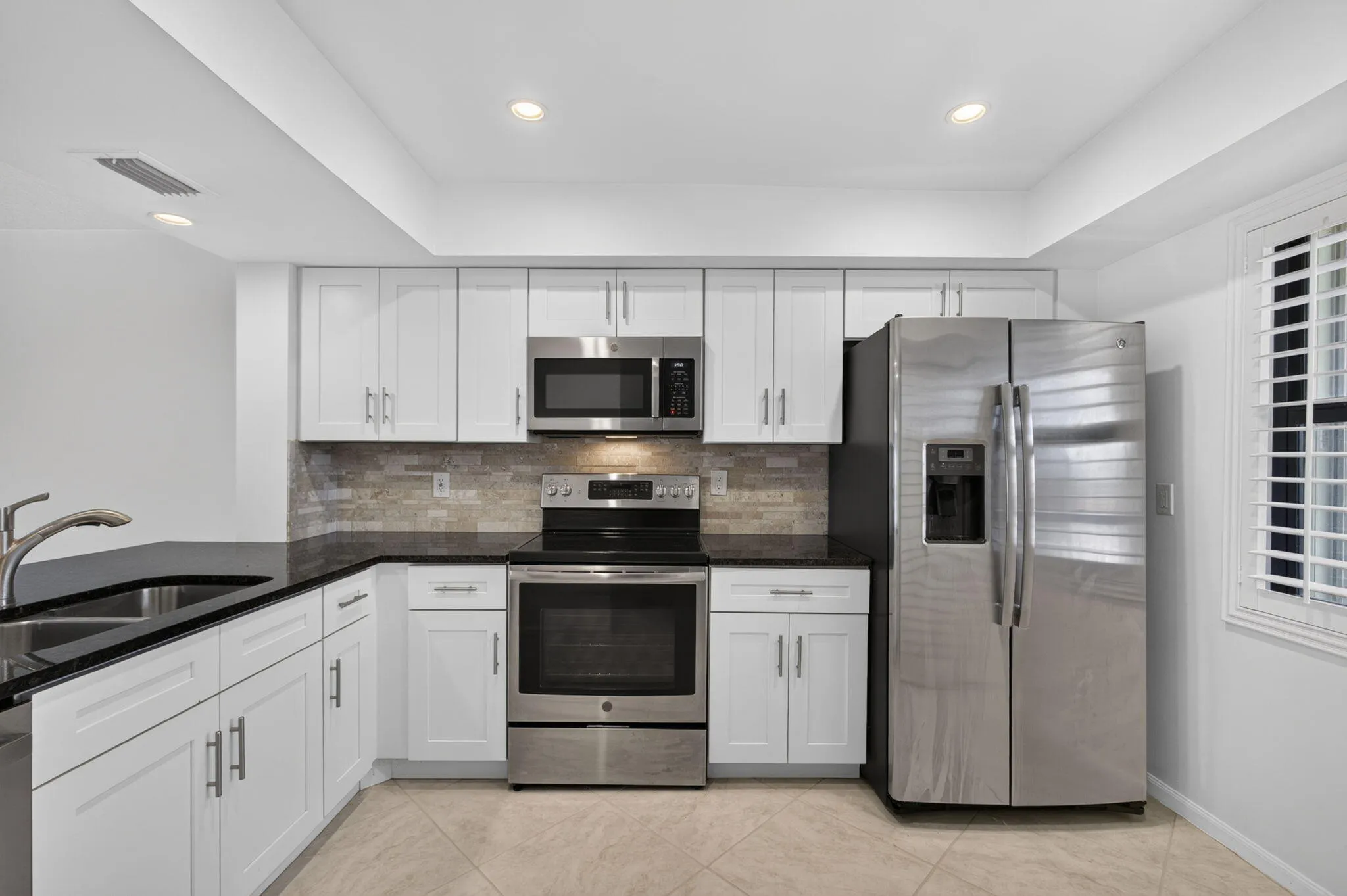 Modern kitchen featuring white shaker cabinetry, stainless steel appliances, black countertops, a neutral-toned tile backsplash, and recessed lighting
