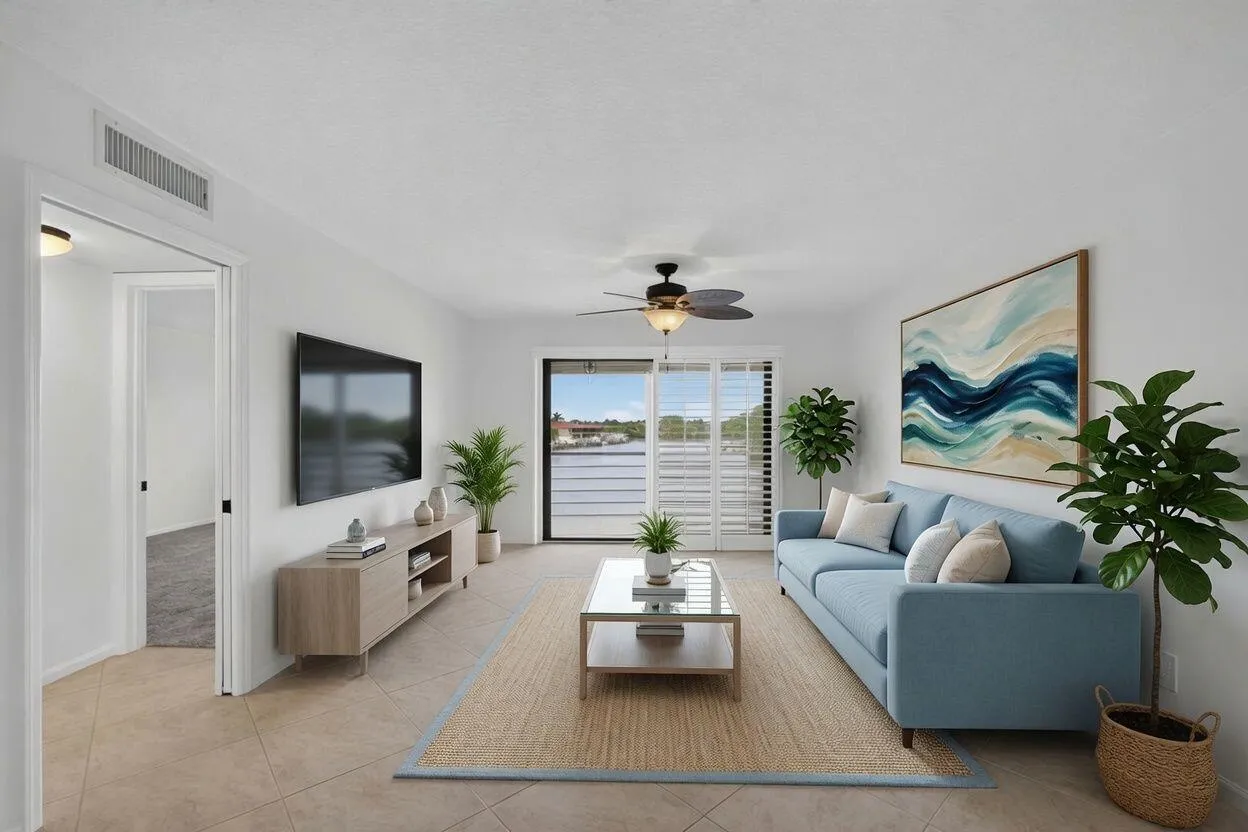 Spacious living area featuring neutral tile flooring, white walls, a ceiling fan, and sliding glass doors with white louvered shutters opening to a scenic water backdrop