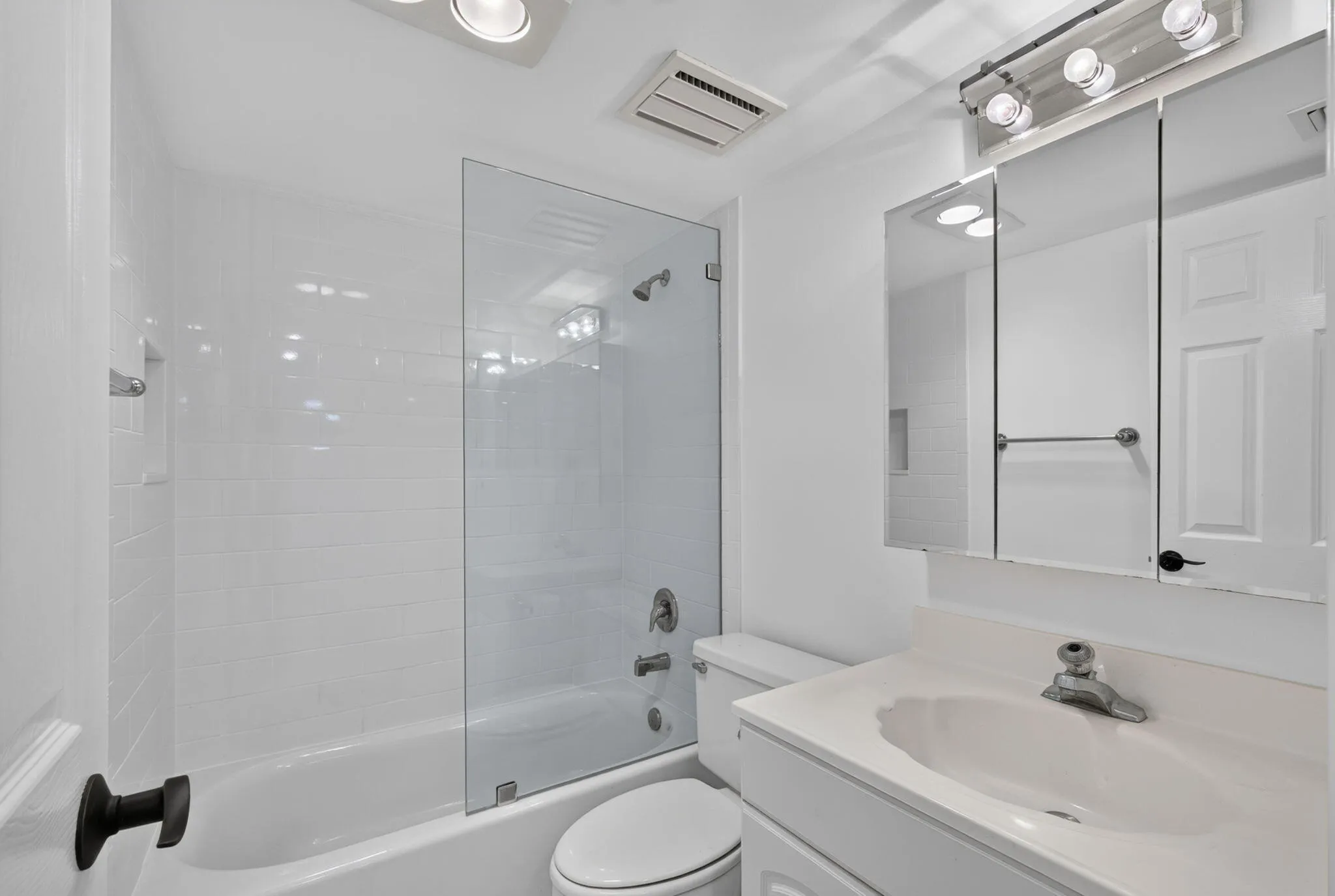 Bright bathroom featuring a white vanity with an integrated sink, a mirrored medicine cabinet with overhead lighting, a white toilet, and a bathtub with white subway tile surround and a clear glass shower panel