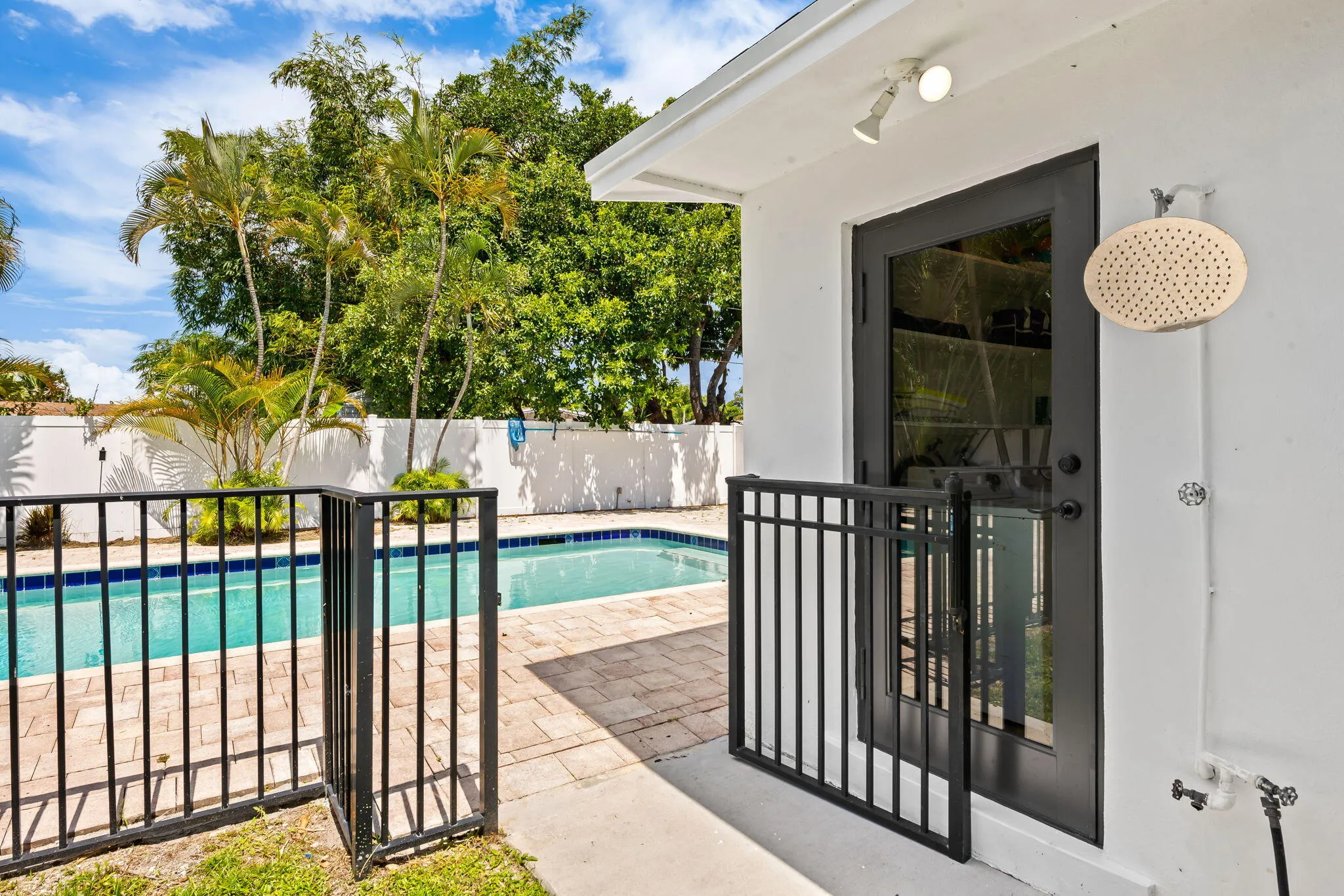Pool area featuring a black aluminum fence with a gate, brick paver decking, an outdoor shower fixture, a black glass-panel door, and mature tropical landscaping