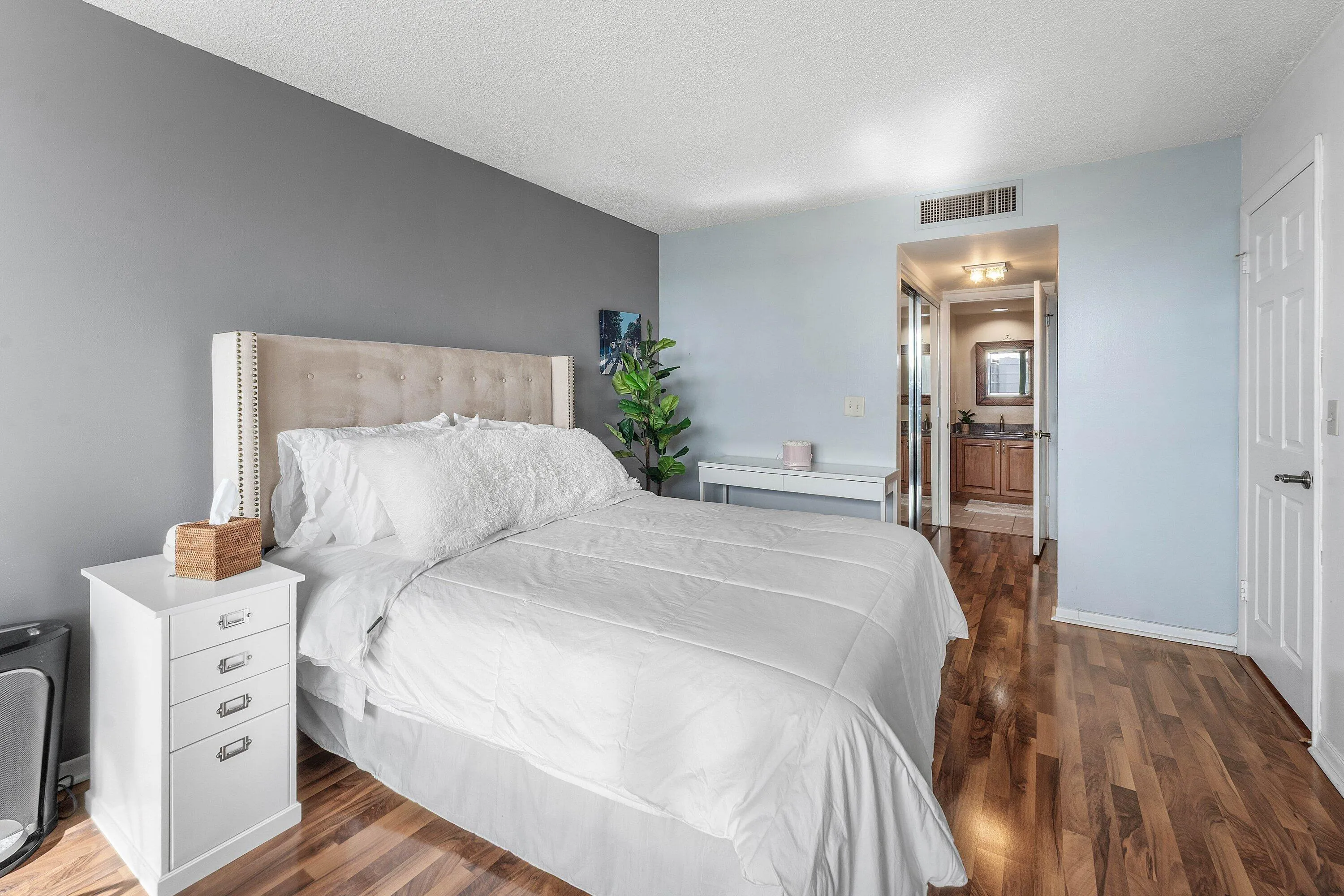 Wood-finish flooring defines this bedroom with a neutral-toned accent wall and a built-in air vent