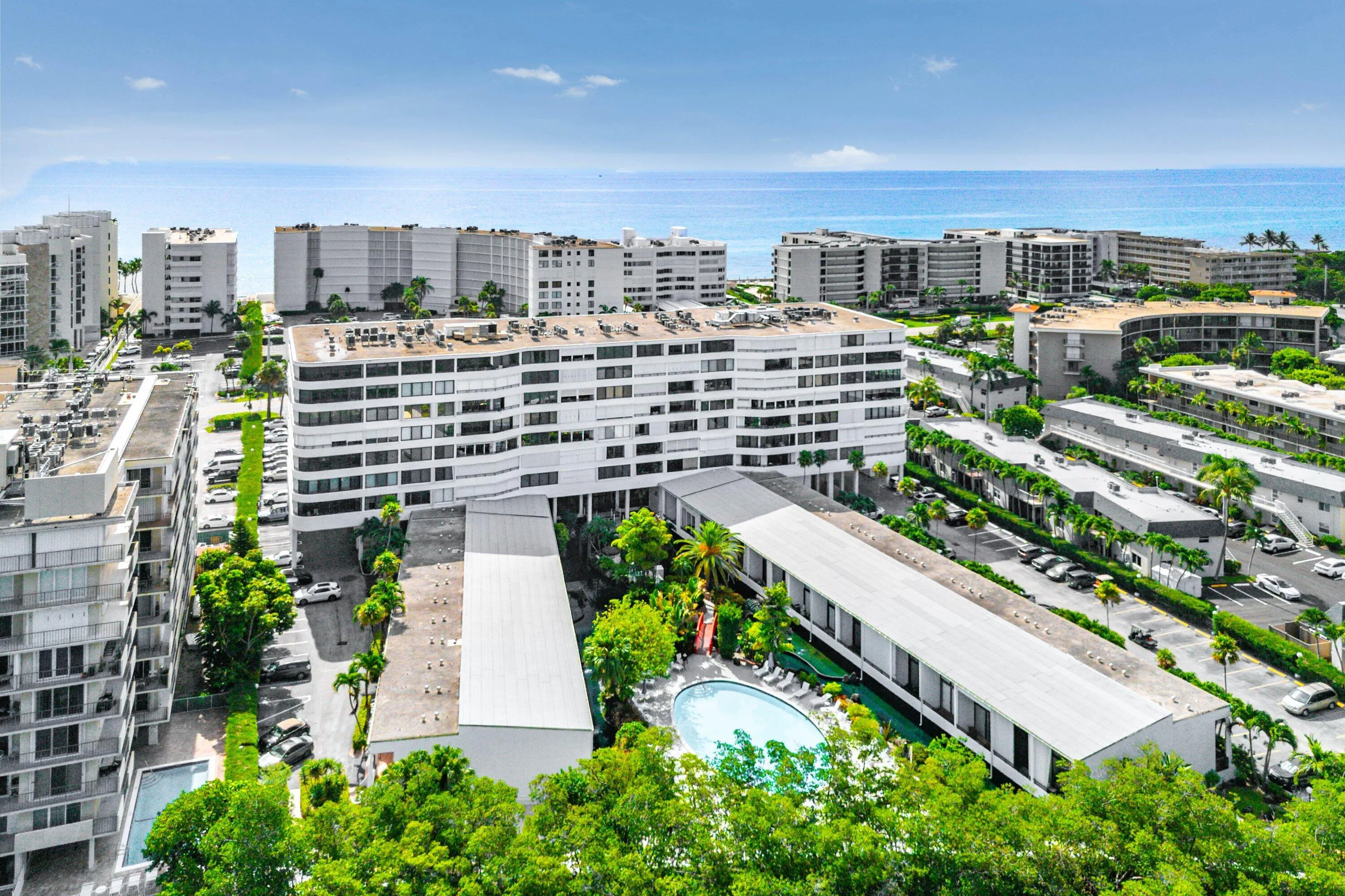 Resort-style pool area with surrounding lush landscaping, expansive paved parking areas, multi-story buildings, and a clear blue sky overhead