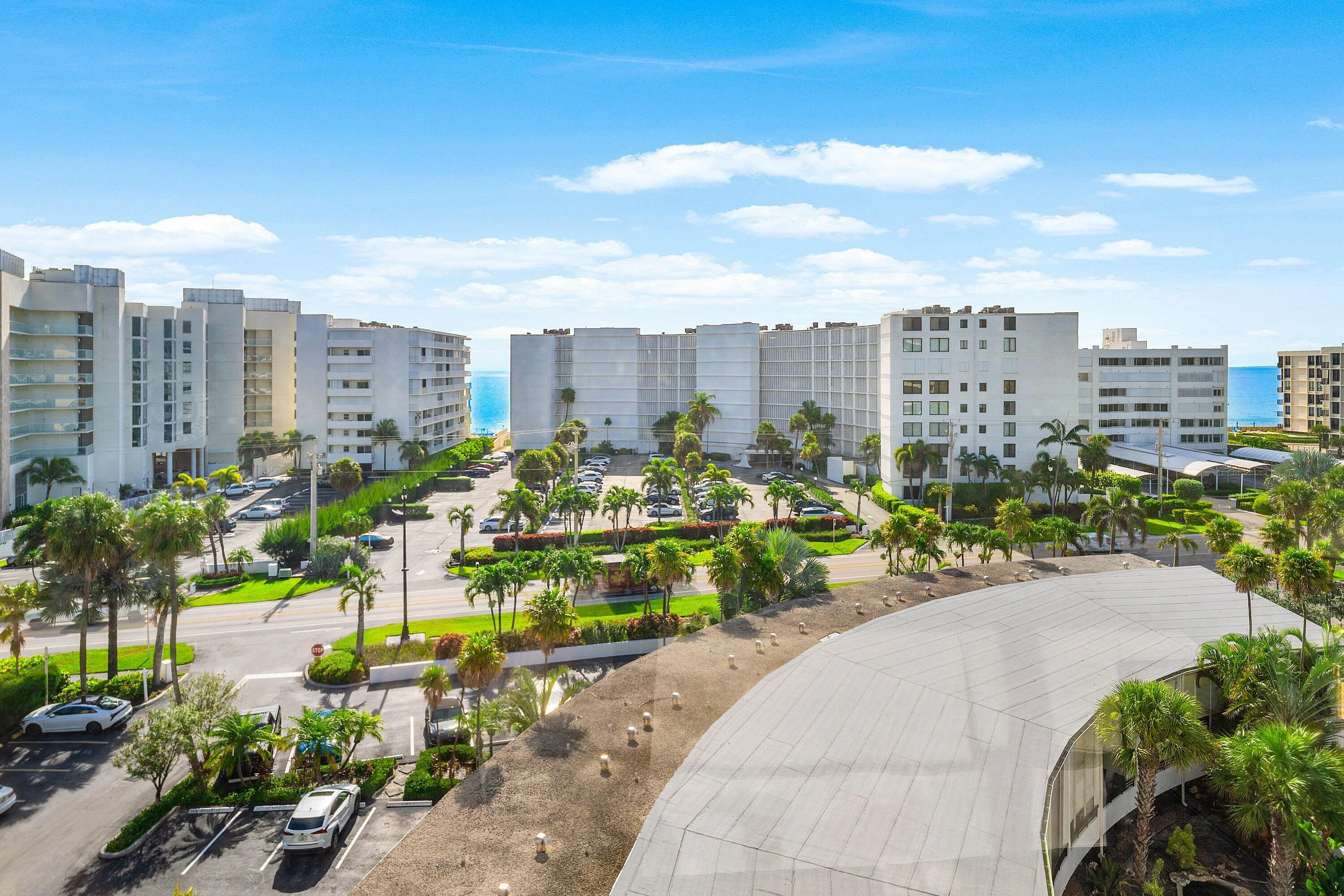 Expansive coastal landscape featuring multiple mid-rise residential buildings with balconies, abundant palm trees, parking areas, and distant ocean water