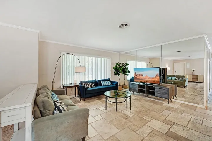 Spacious living area featuring large format tile flooring, a mirrored accent wall, crown molding, and a wide window with vertical blinds