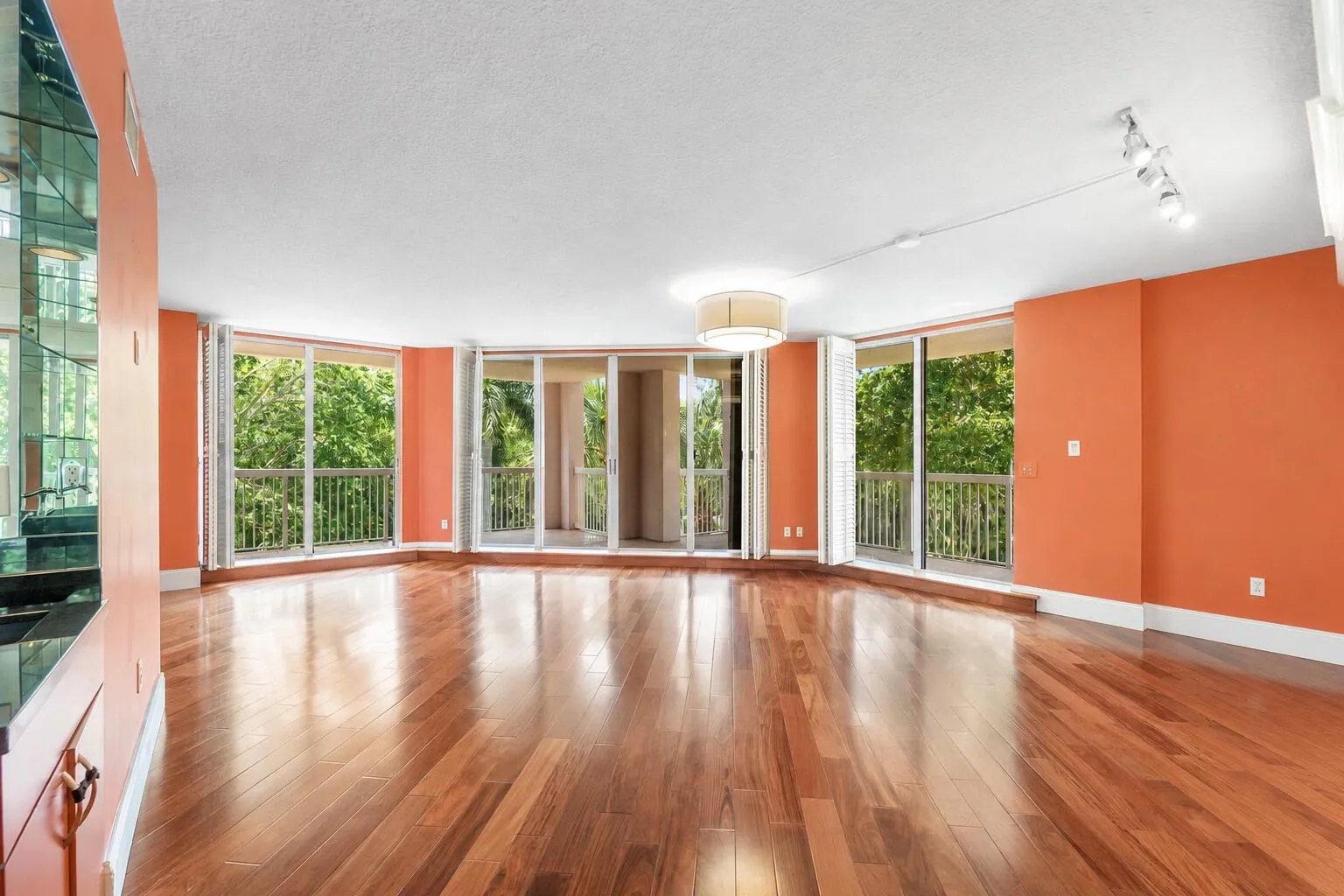 Expansive living area featuring rich wood flooring throughout, multiple sliding glass doors opening to outdoor balconies, a contemporary drum chandelier, track lighting, and a built-in mirrored wet bar with a dark countertop