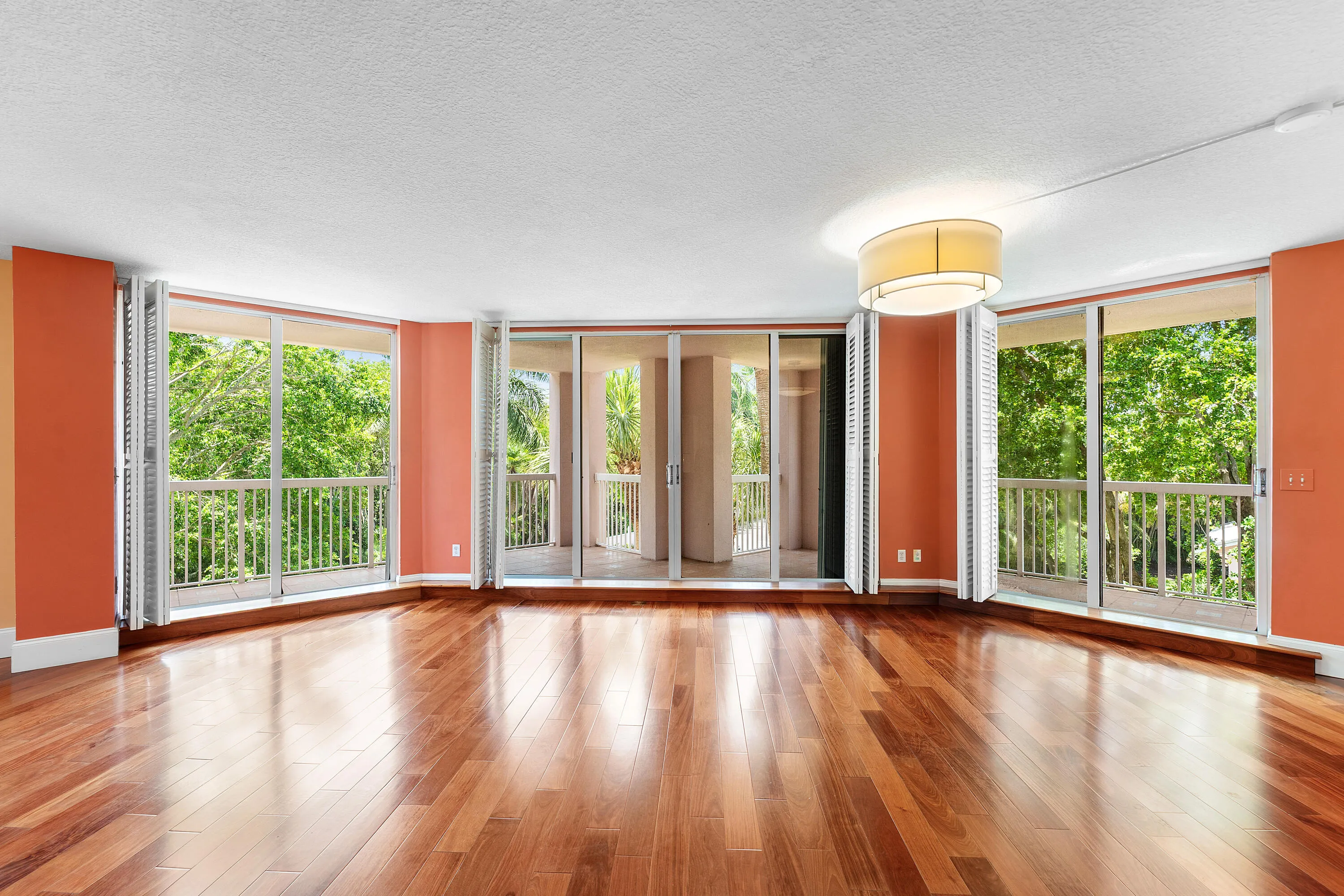 Spacious room featuring wood-finish flooring, a contemporary drum pendant light fixture, and multiple glass sliding doors with white louvered shutters