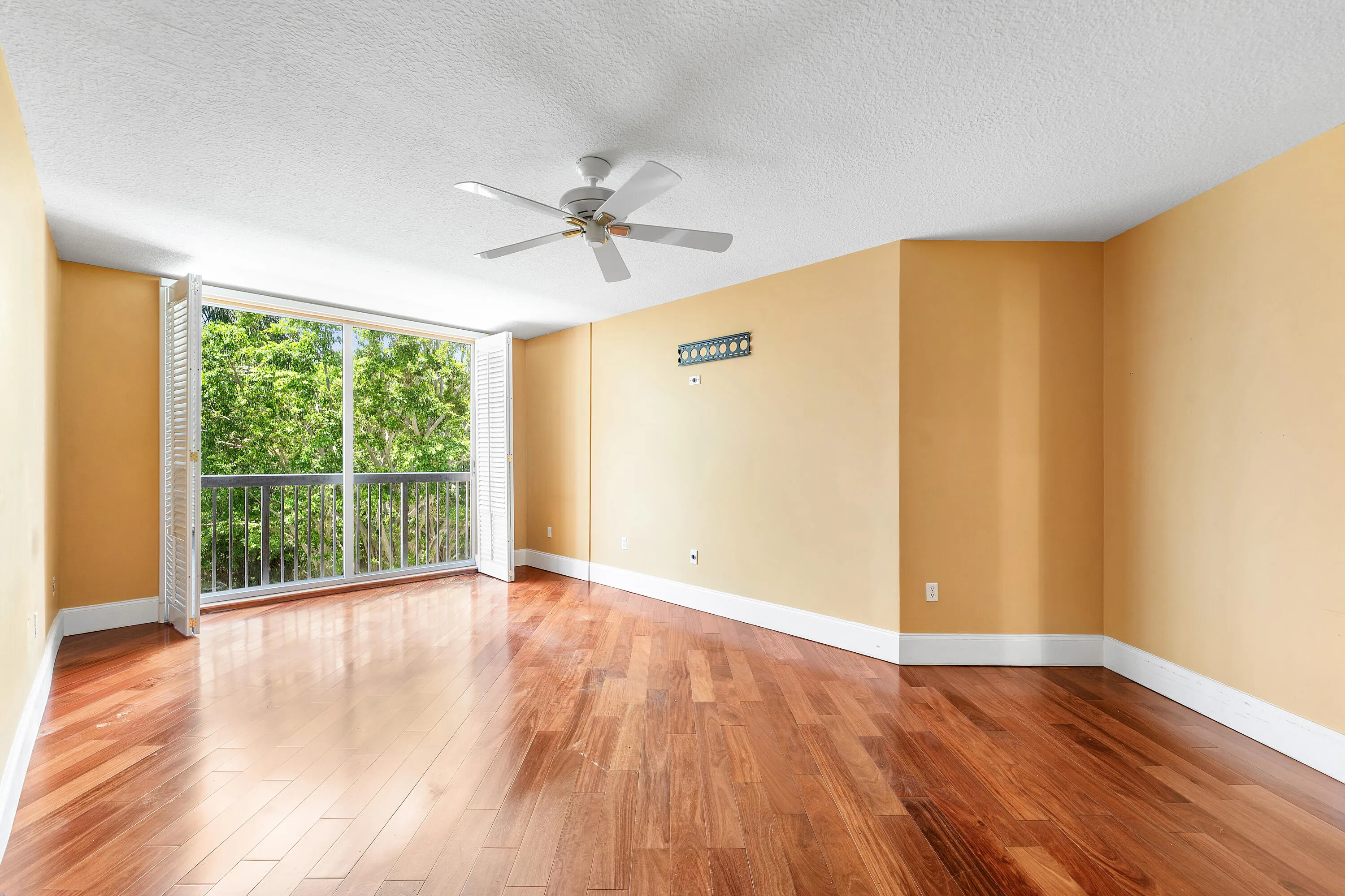 Spacious room featuring wood-finish flooring, a ceiling fan, and a large window with white louvered shutters