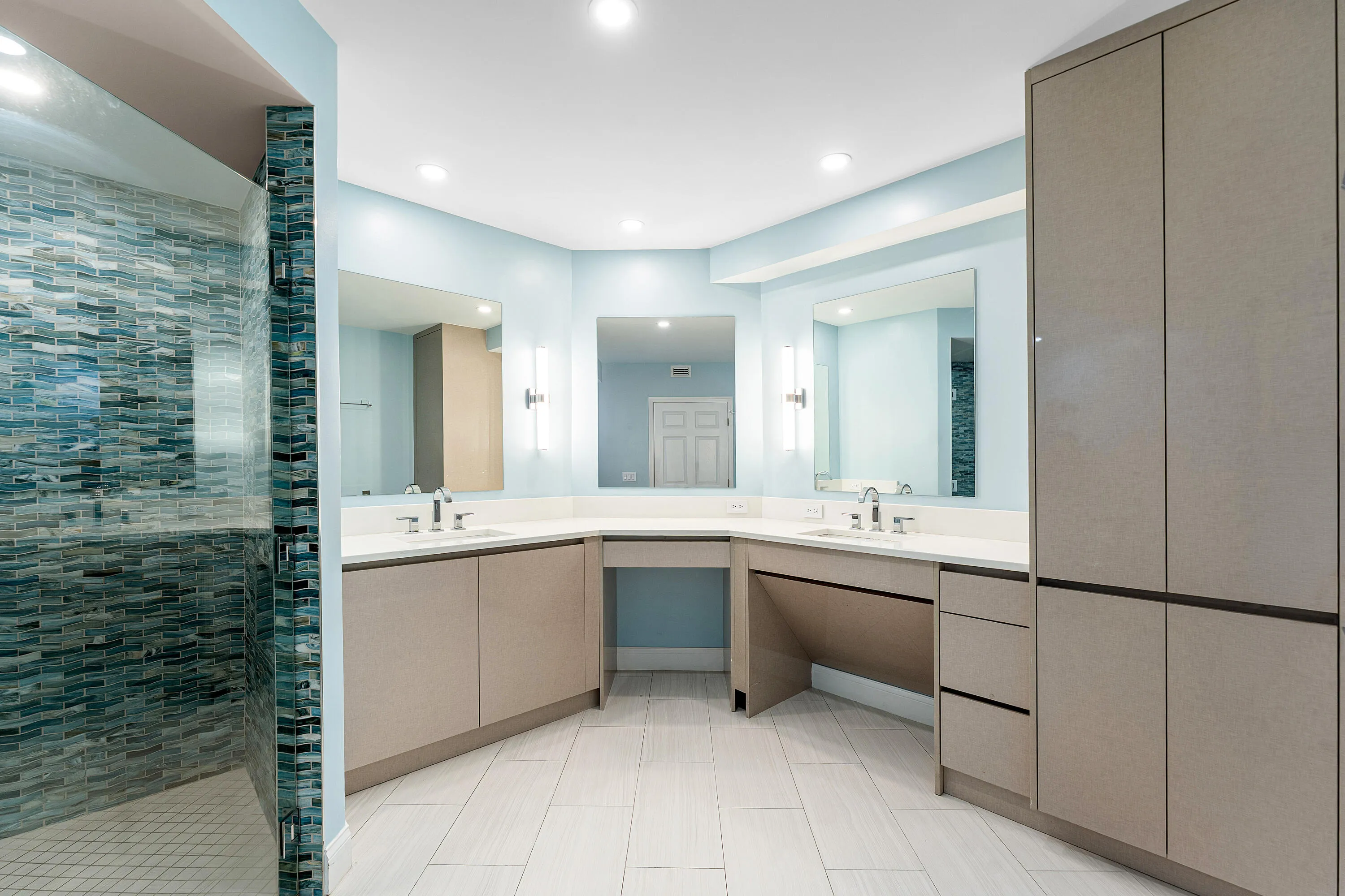 Spacious bathroom featuring a glass-enclosed shower with blue mosaic tile, dual vanity sinks, a built-in makeup vanity, and extensive built-in storage cabinetry