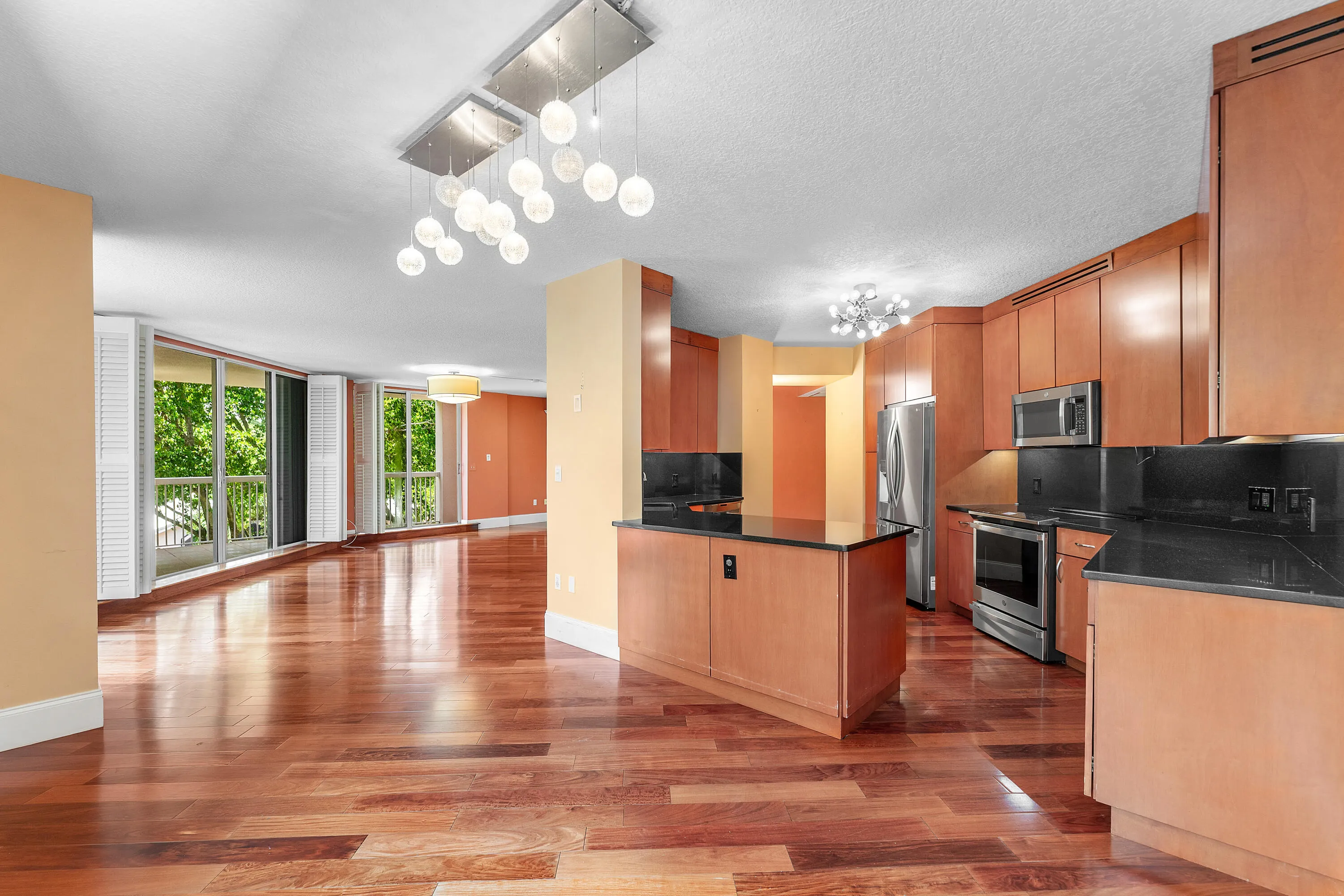 Open-concept living space featuring wood-finish flooring, a full kitchen with light wood cabinetry and dark countertops, stainless steel appliances, and abundant natural light from large windows with exterior foliage