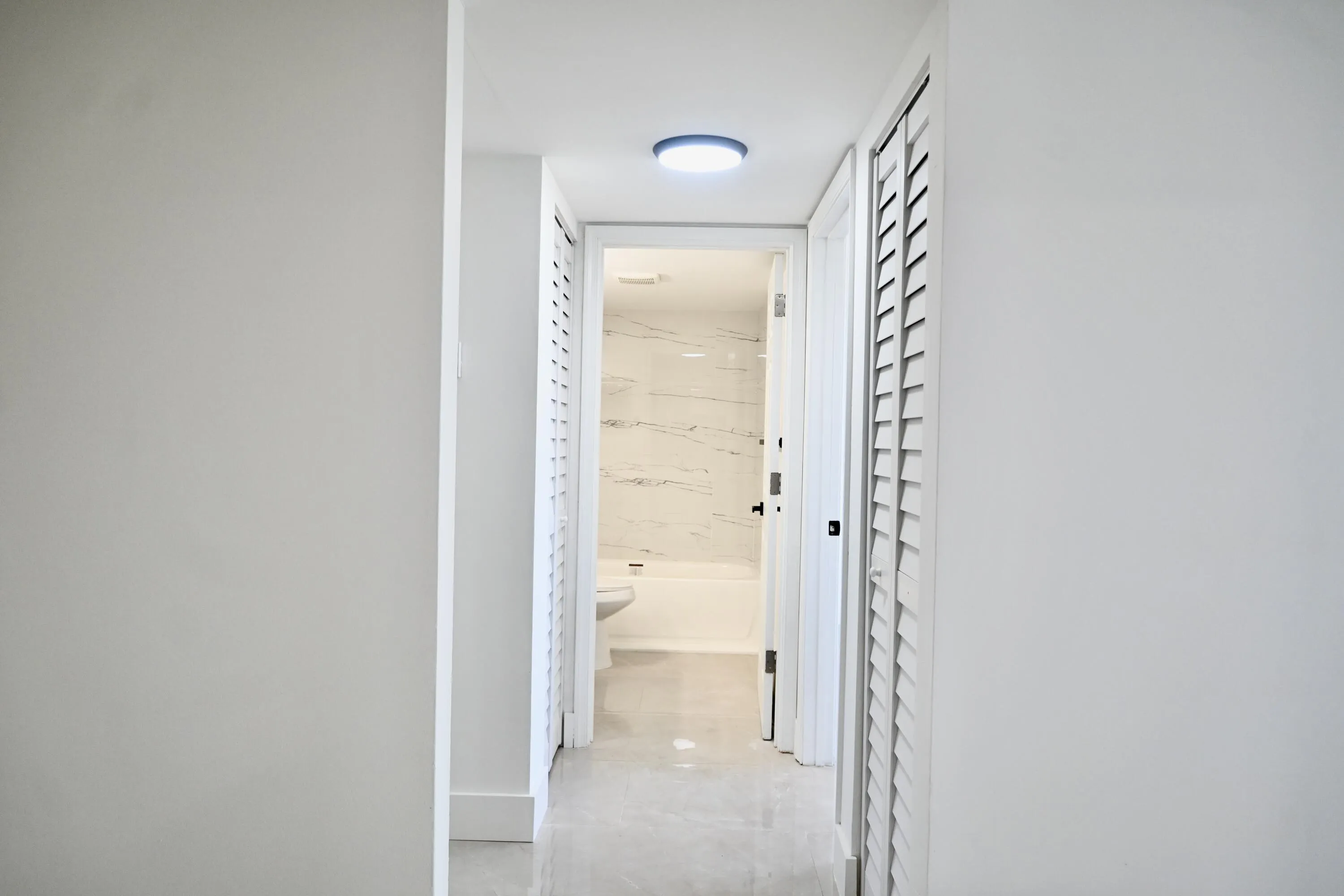 Hallway with recessed lighting, louvered closet doors, and light-tone flooring leading to a bathroom featuring a white bathtub and marble-patterned wall tiles