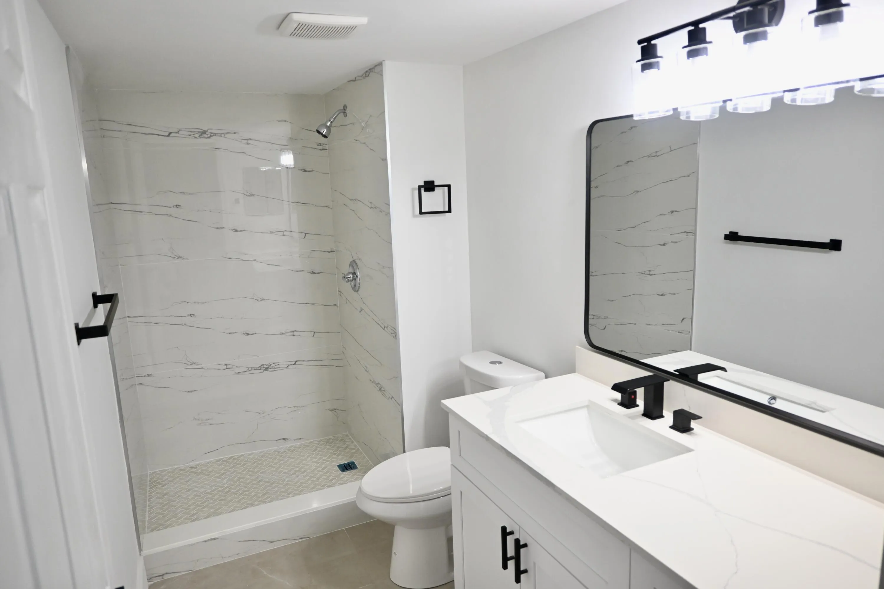Modern bathroom featuring a walk-in shower with white veined tile surround, a white vanity with integrated sink, matte black fixtures, a rectangular vanity mirror, and overhead lighting