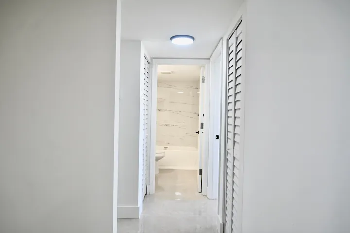 Hallway with recessed lighting, louvered closet doors, and light-tone flooring leading to a bathroom featuring a white bathtub and marble-patterned wall tiles