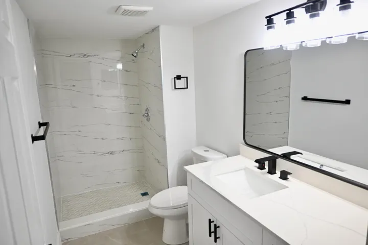 Modern bathroom featuring a walk-in shower with white veined tile surround, a white vanity with integrated sink, matte black fixtures, a rectangular vanity mirror, and overhead lighting
