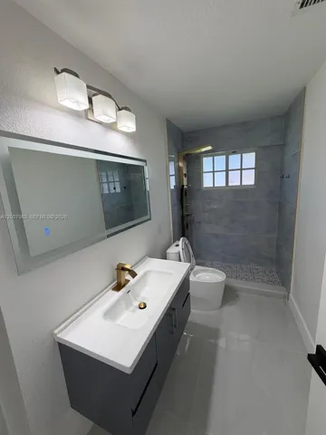 Floating vanity with integrated sink, brass-finish faucet, illuminated mirror, and a walk-in shower with tiled walls