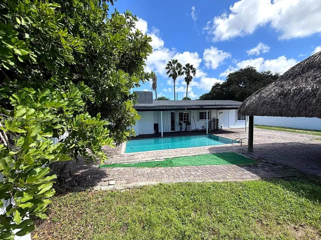In-ground swimming pool with brick paver surround, adjacent to a structured tiki-style hut, and a white exterior residence